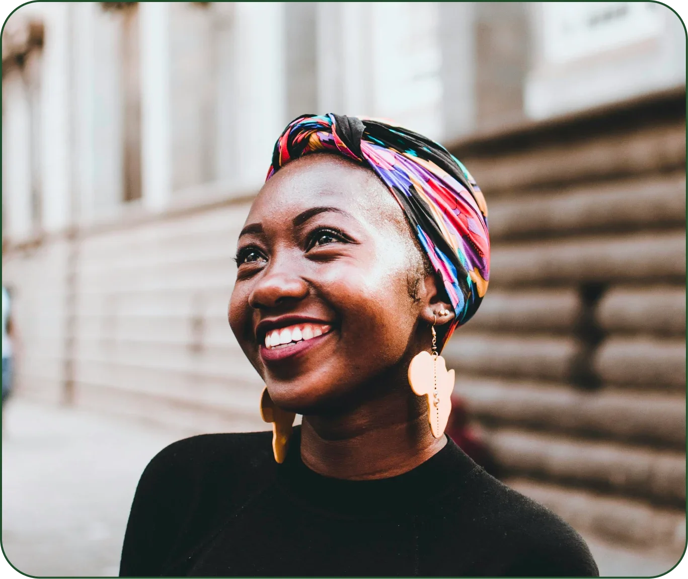 Close-up of smiling African American woman with colorful head wrap, large earrings, and black top, standing outdoors with blurred wooden background.