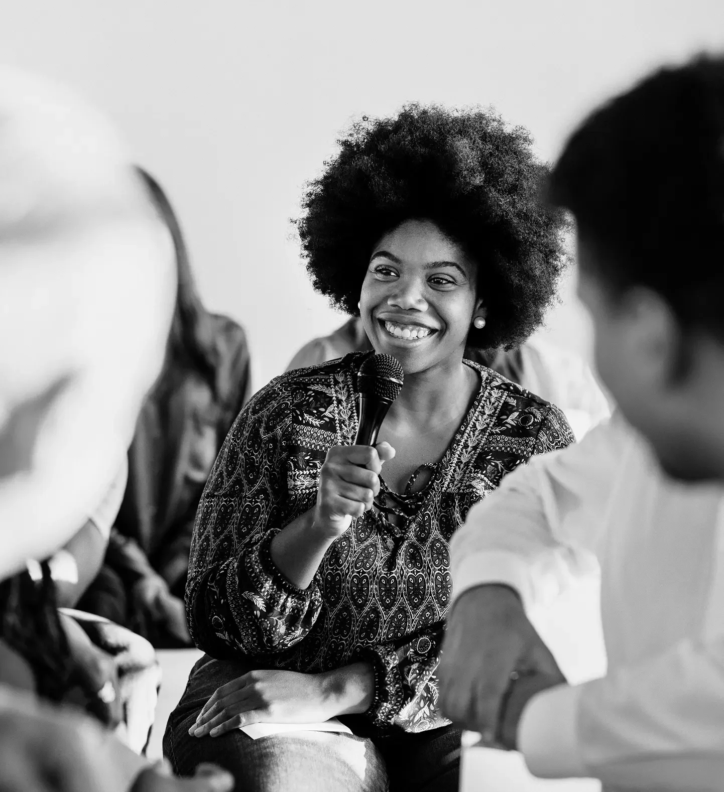 Black and white photo of a woman with an afro hairstyle, smiling and holding a microphone, engaging with a group of people in a discussion or interview setting.