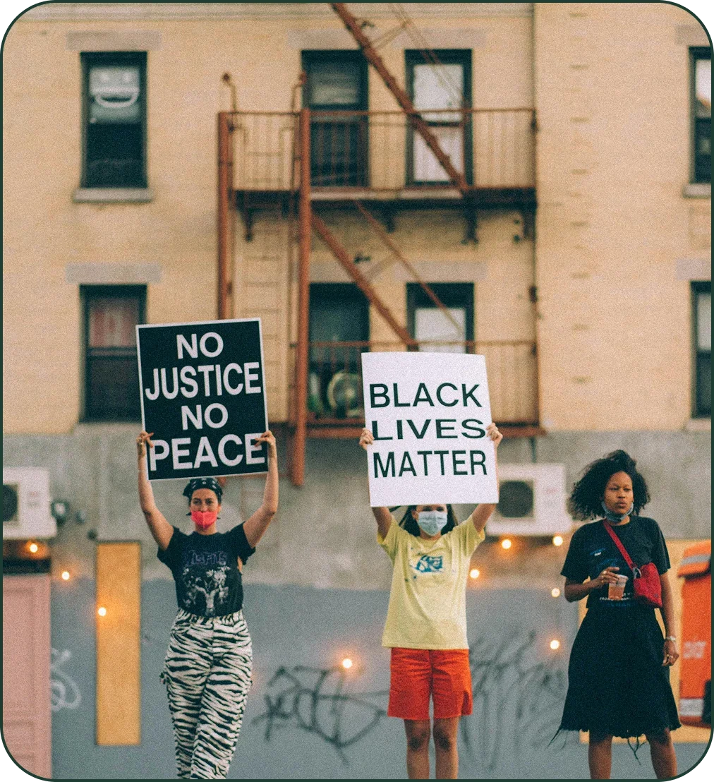 Three women protesting on the street, holding signs that read "NO JUSTICE NO PEACE" and "BLACK LIVES MATTER", with a beige building and fire escape in the background