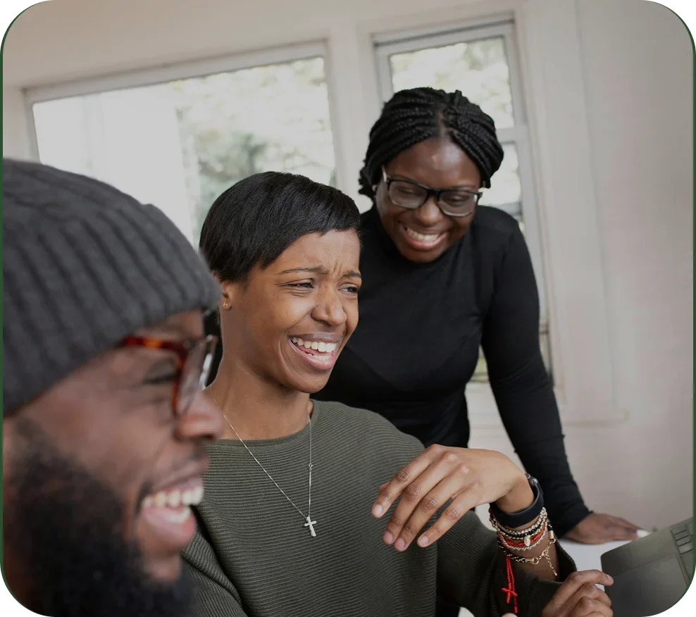 Three friends laughing and looking at a tablet in a bright room with large windows.