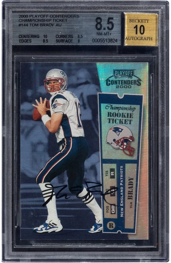 A football trading card of Tom Brady in a Patriots uniform, inside a protective case. The card is labeled as a Rookie Ticket from the 2000 Playoff Contenders. It features Tom Brady in a passing stance and an autograph signature at the bottom. The case has a grade of 8.5 out of 10 and includes a Beckett autograph grade of 10.