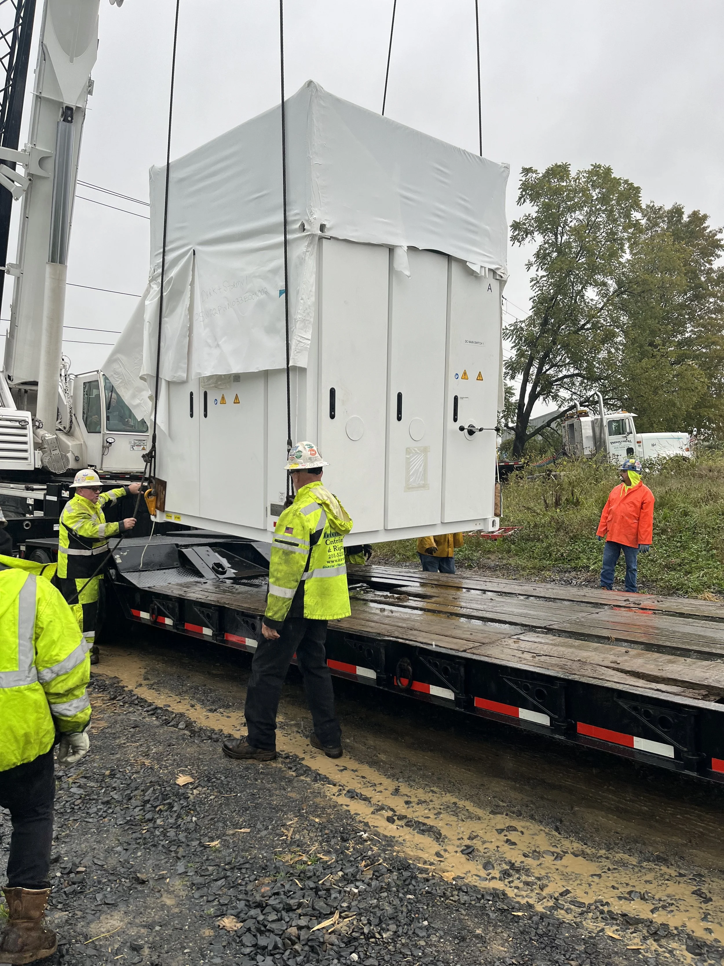 Workers in high-visibility clothing and helmets are overseeing the installation of a large, white electrical transformer on a flatbed truck at a construction site on a cloudy day. The ground is muddy with gravel and fallen leaves.