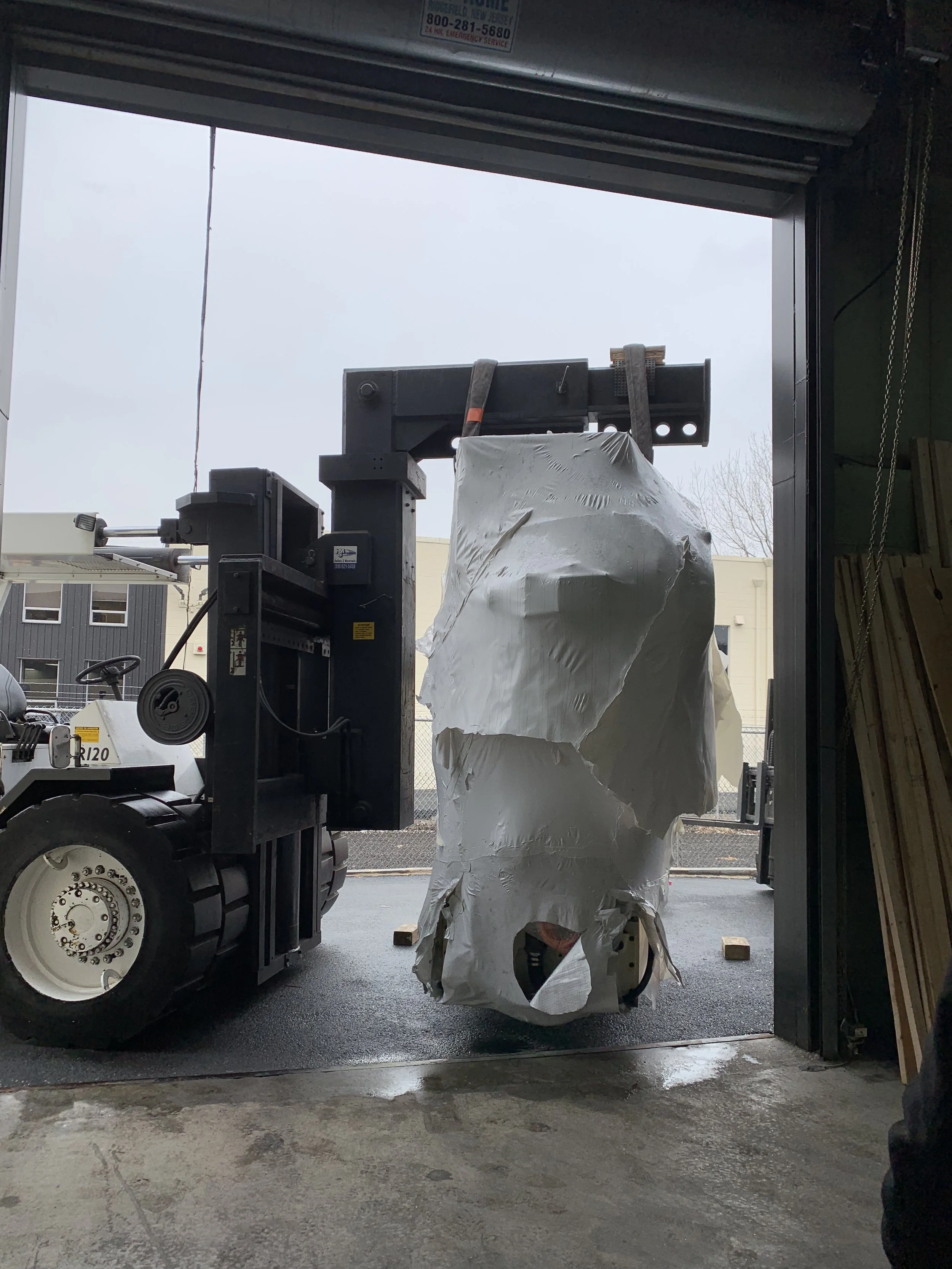 A forklift is lifting a large, white, crumpled bag or sheet in a warehouse or industrial setting, with a view from inside a garage or loading bay.