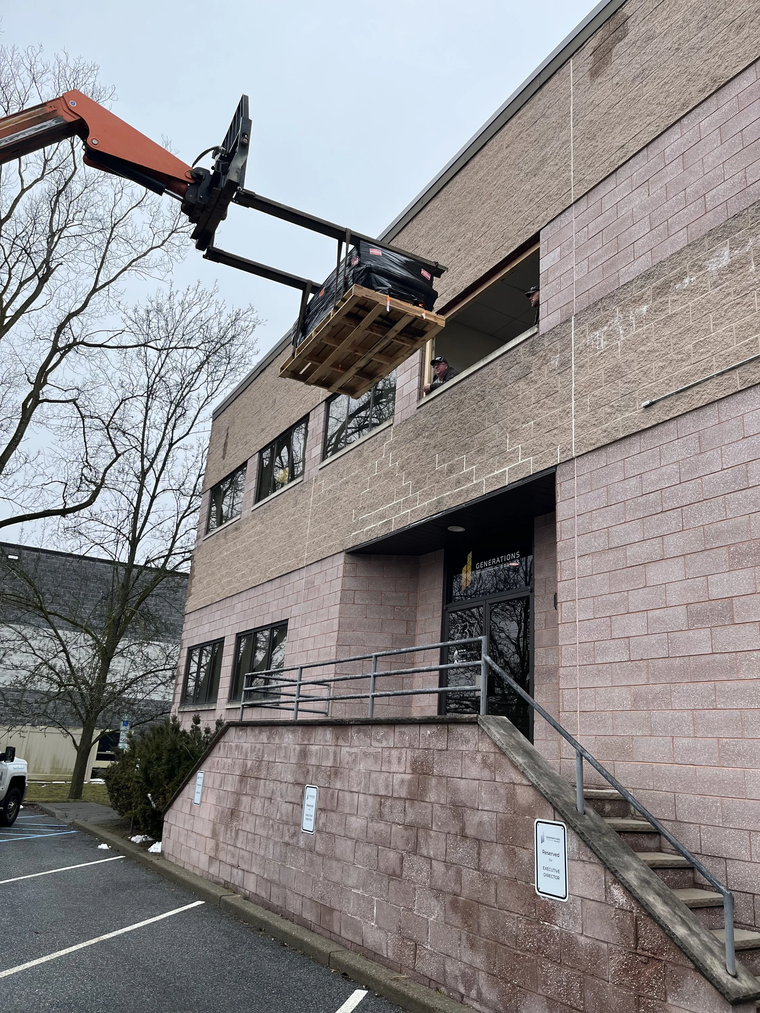 A building with a balcony being worked on with a crane lifting materials and workers observing. The building has a brick and concrete exterior, some parking spaces, and reserved signs for executives and the director.