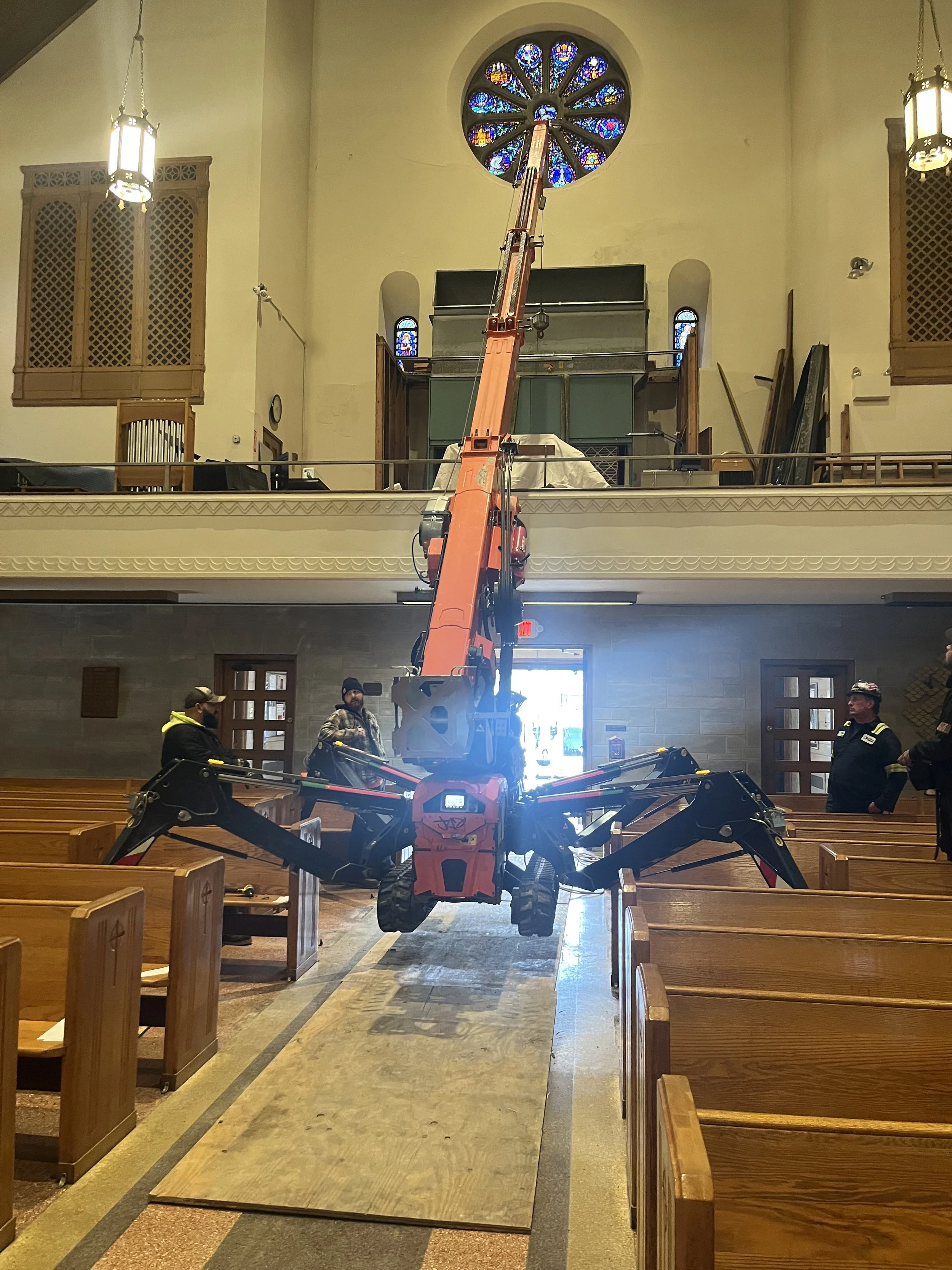 Inside a church with wooden pews, a large stained glass window, and a large orange crane in the center adjusting or installing something near the ceiling.
