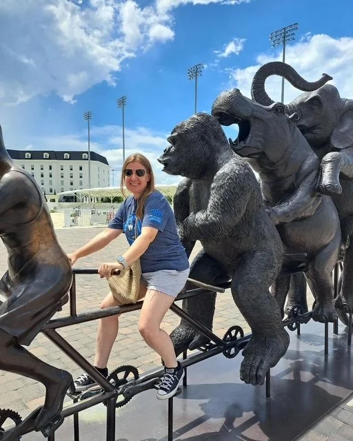A woman with sunglasses smiling while riding a sculpture of a horse, passing by a bear, a lion, and an elephant, outdoors on a sunny day.