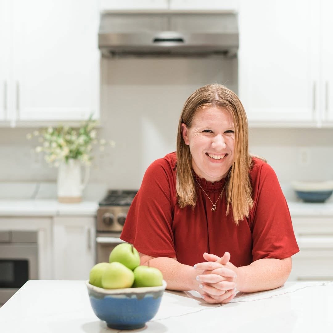 A woman with shoulder-length brown hair, wearing a red top, smiling and leaning on a white kitchen counter. There is a blue bowl with green apples in front of her. The background shows a kitchen with white cabinets, a stove, and a vase with flowers.