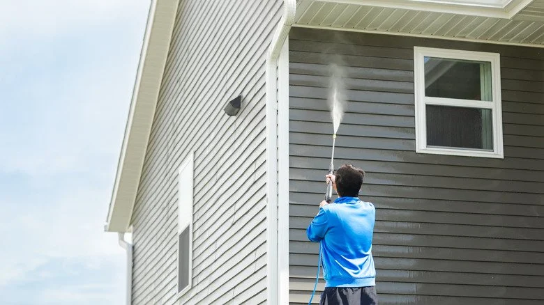 A man in a blue jacket using a pressure washer to clean the side of a house with gray siding.
