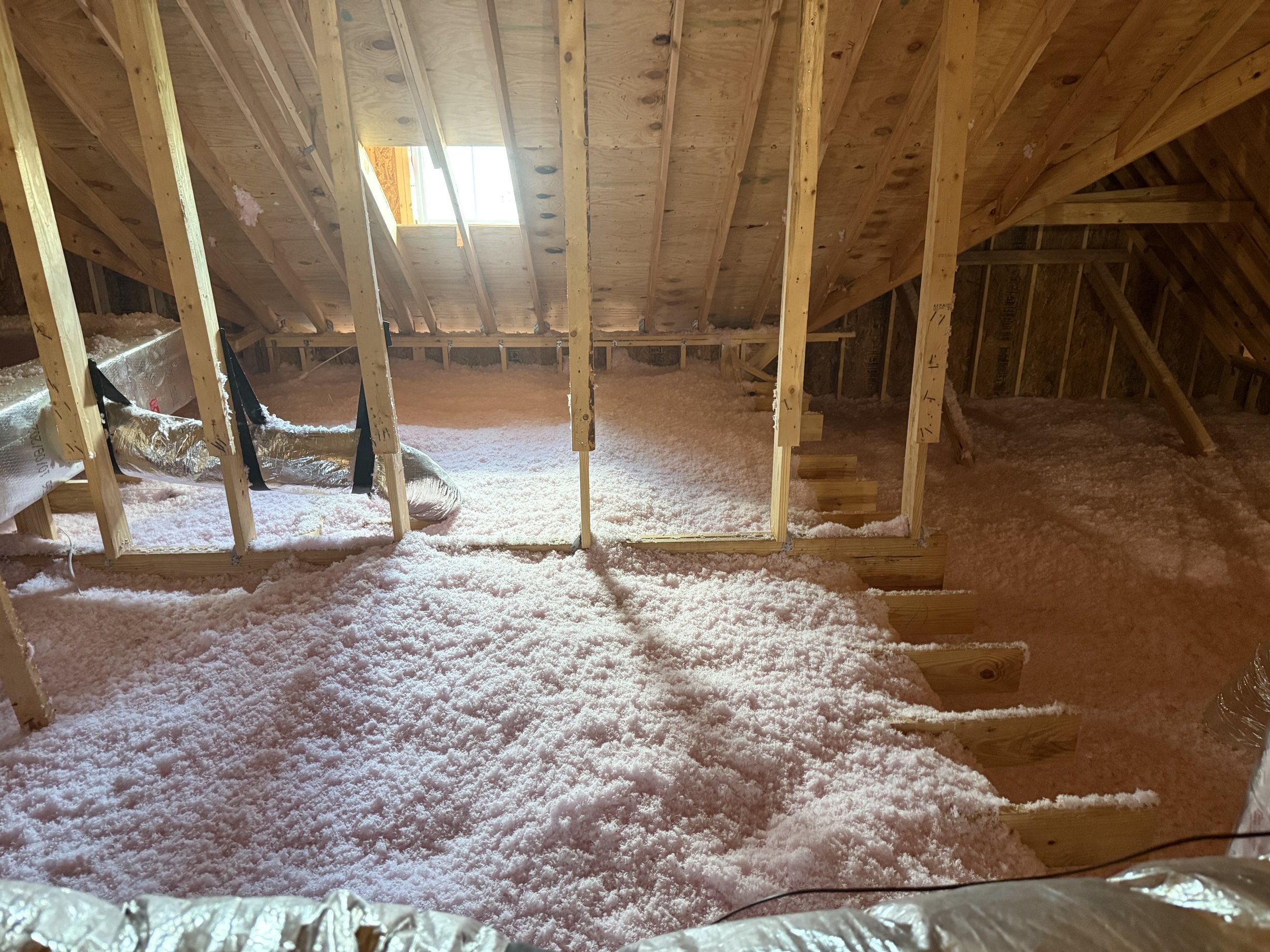 Attic with exposed wooden framing, insulation on floor, and a small window letting in light.
