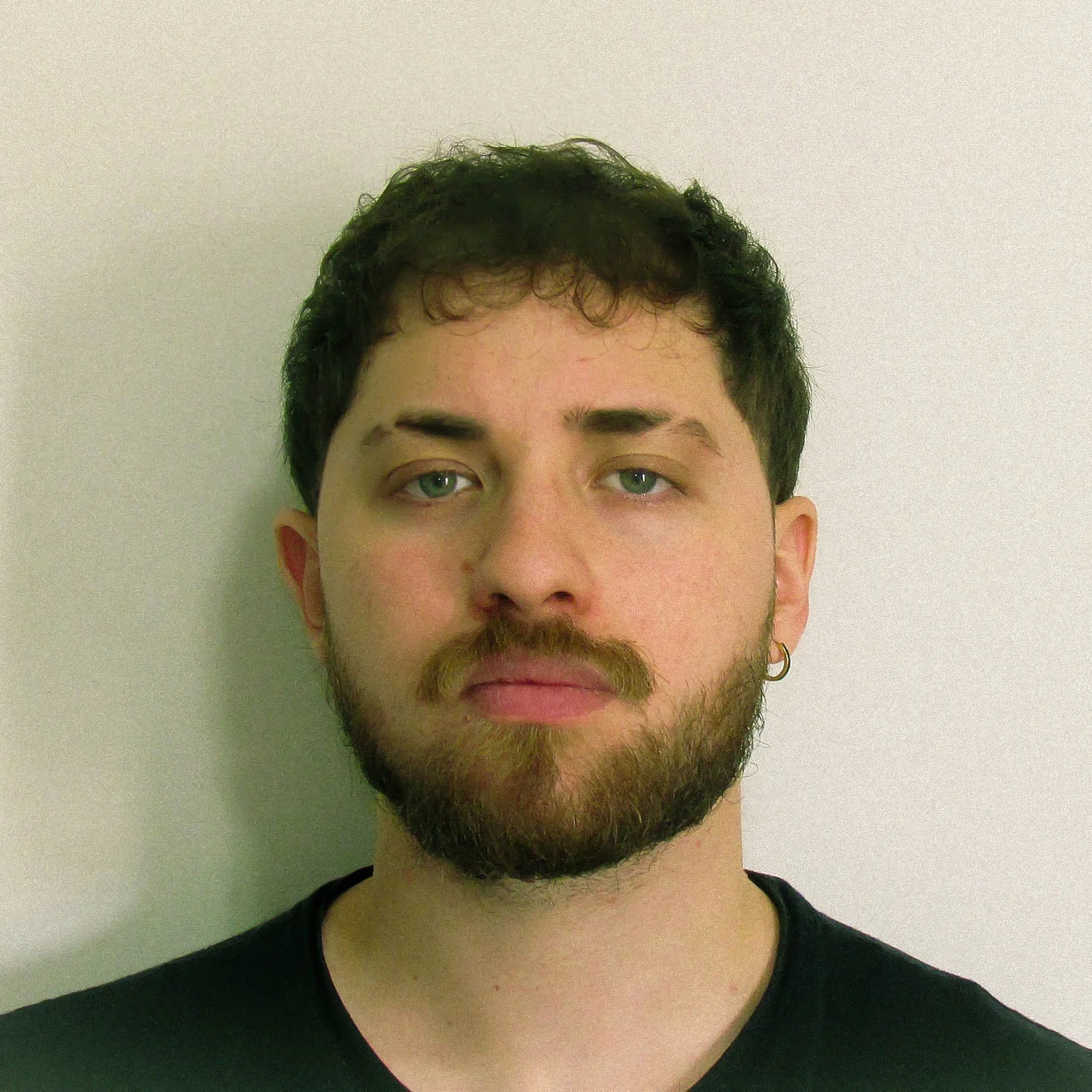 Retrato de um homem com barba, olhos verdes e cabelo escuro encaracolado, vestindo uma camiseta preta, contra um fundo claro