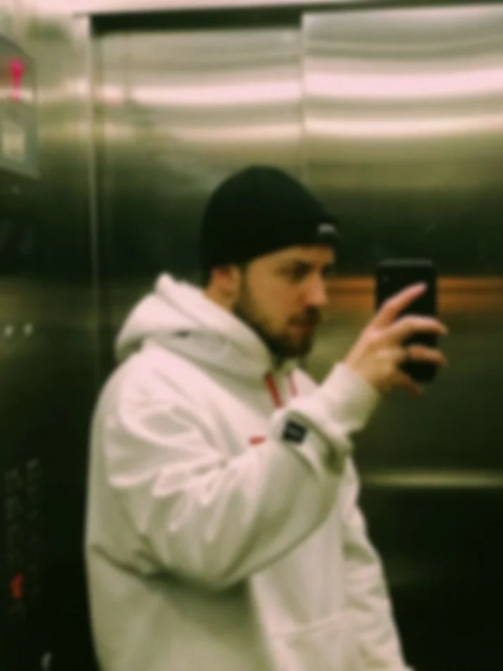 Homem com barba, usando blusa branca e gorro preto, tirando selfie em um elevador de espelho