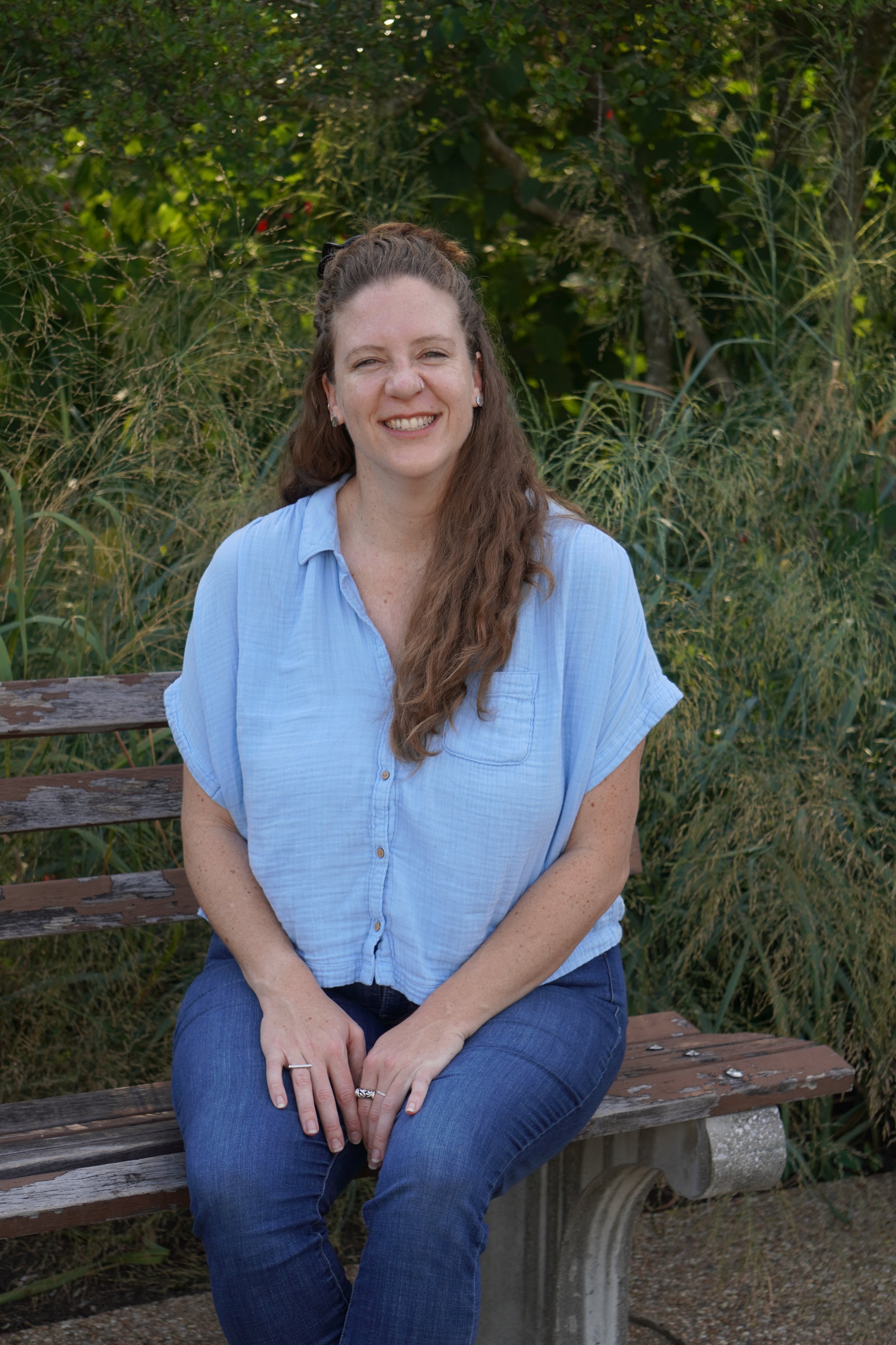 A woman with long curly hair, smiling, wearing a light blue button-up shirt and jeans, sitting on a wooden park bench outdoors with green foliage in the background.