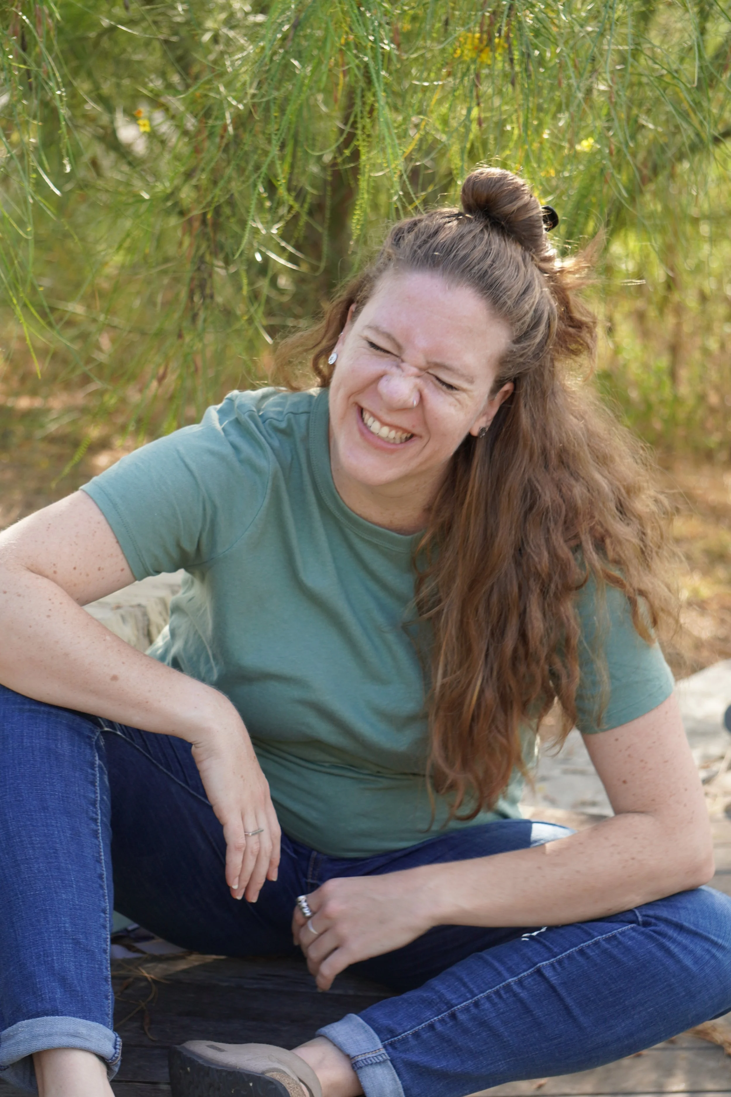 A woman with long hair tied in a half bun, smiling and squinting, sitting outdoors near green trees, wearing a green t-shirt and blue jeans.