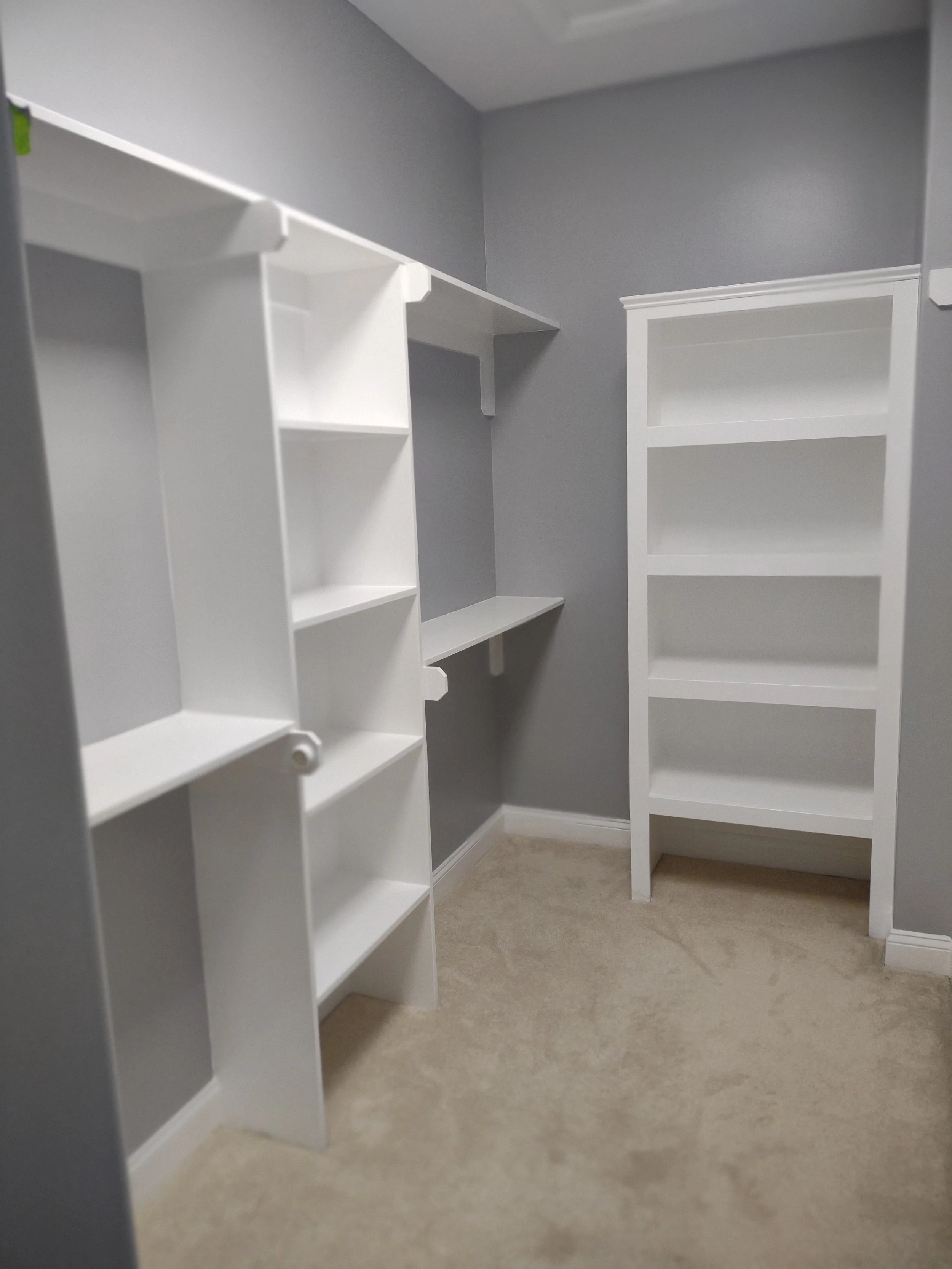 A walk-in closet with white shelving units and beige carpeted flooring.