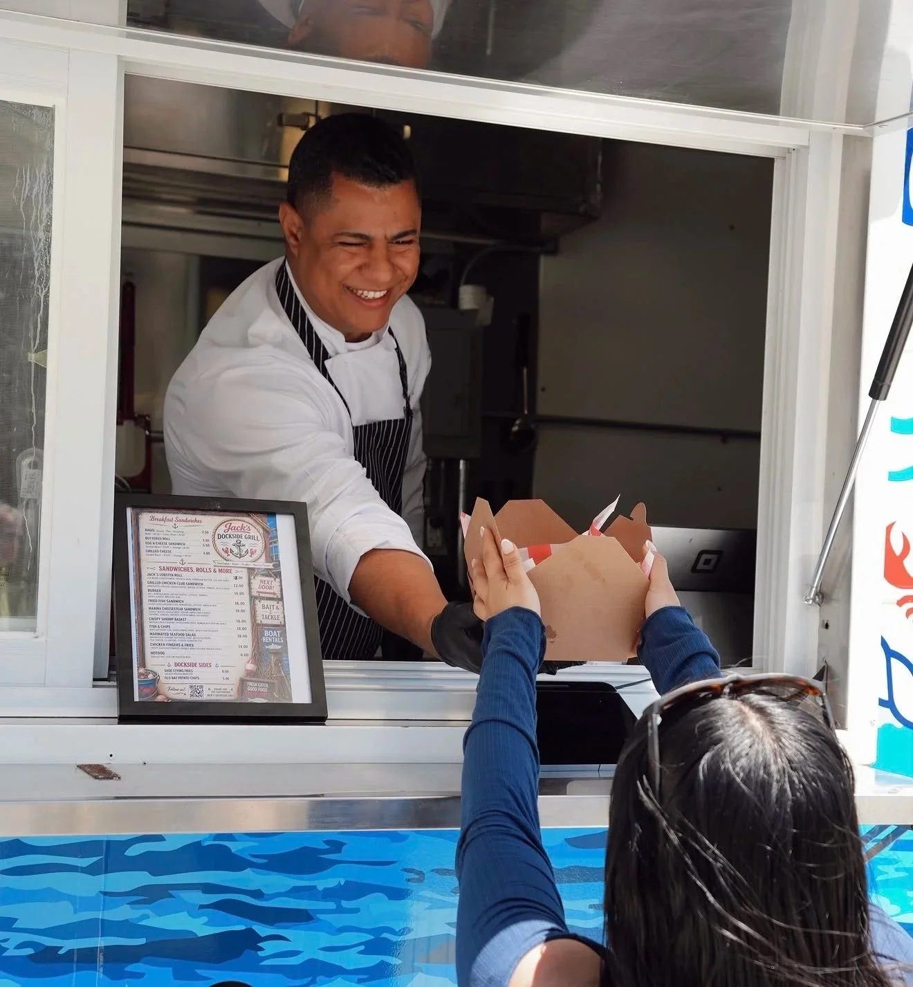 A man in a white shirt and striped apron is handing a takeout container to a customer at a food truck. The customer is wearing a blue jacket and sunglasses on her head, reaching out to receive the food.
