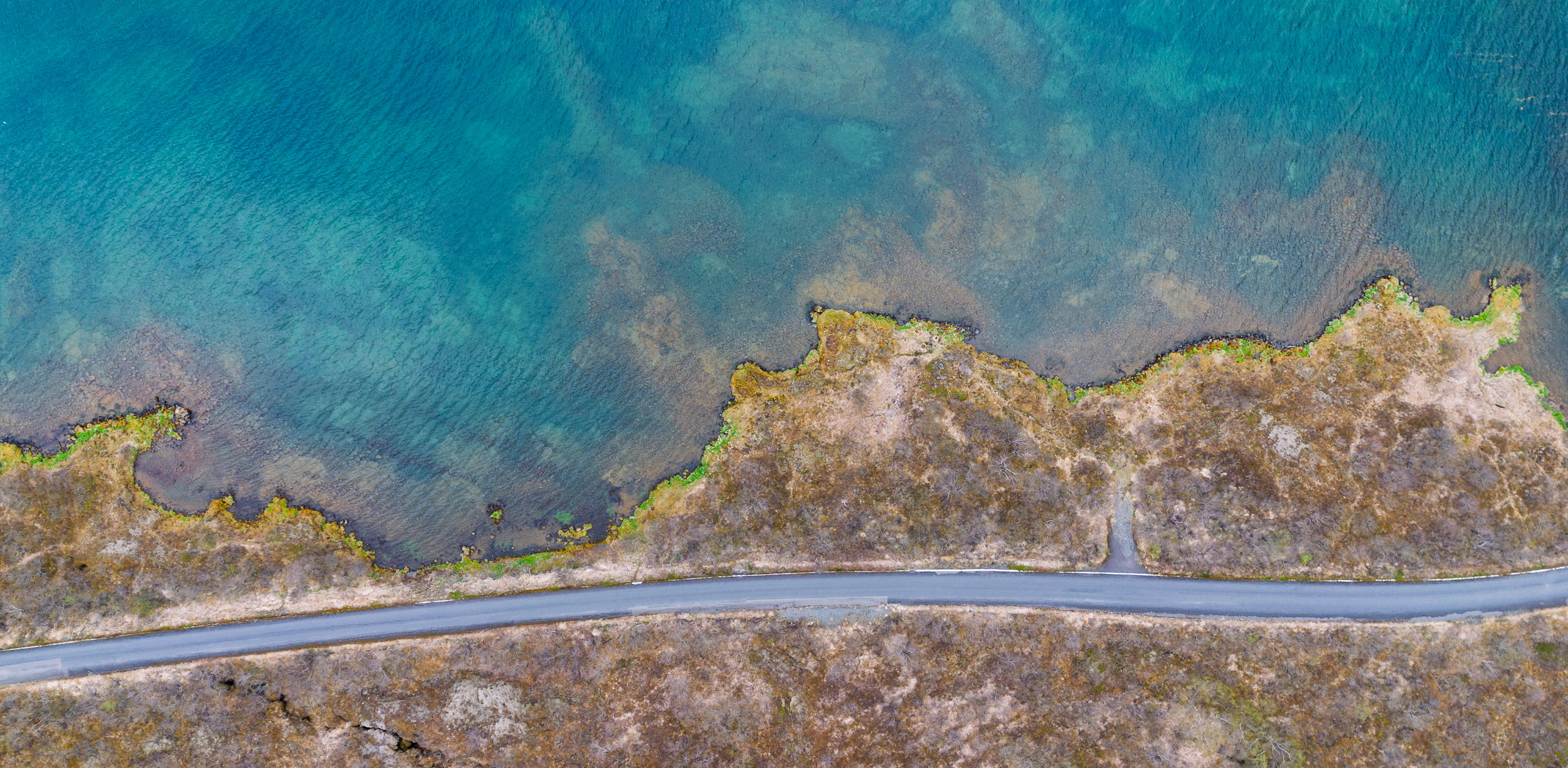 Aerial view of a coastline in Iceland with clear blue water and a winding road along the rocky shore.