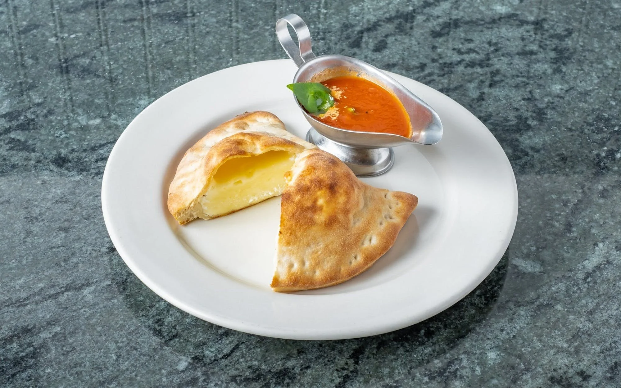 A white plate with a folded stuffed naan bread, a slice exposing melted cheese inside, and a small silver gravy boat with tomato-based curry topped with a green cilantro leaf.