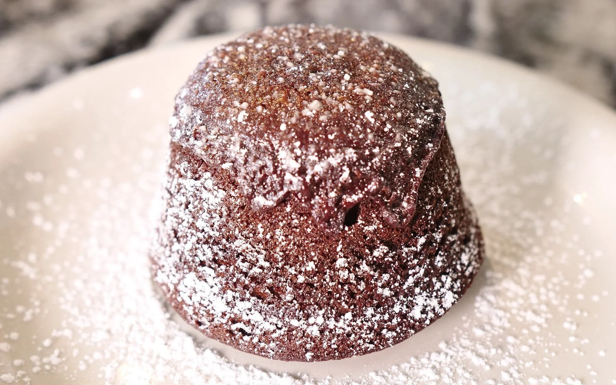 Close-up of a chocolate muffin dusted with powdered sugar, on a white plate with powdered sugar scattered around.