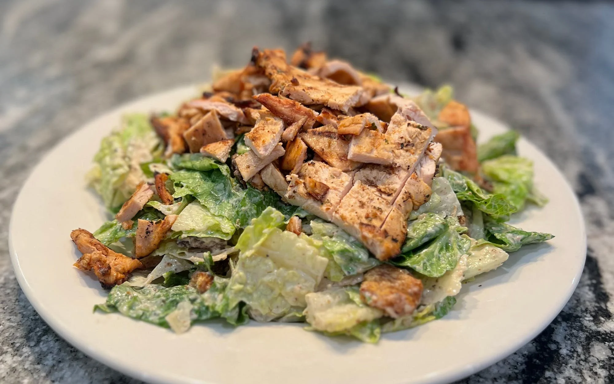 A plate of Caesar salad topped with grilled chicken pieces on a gray countertop.
