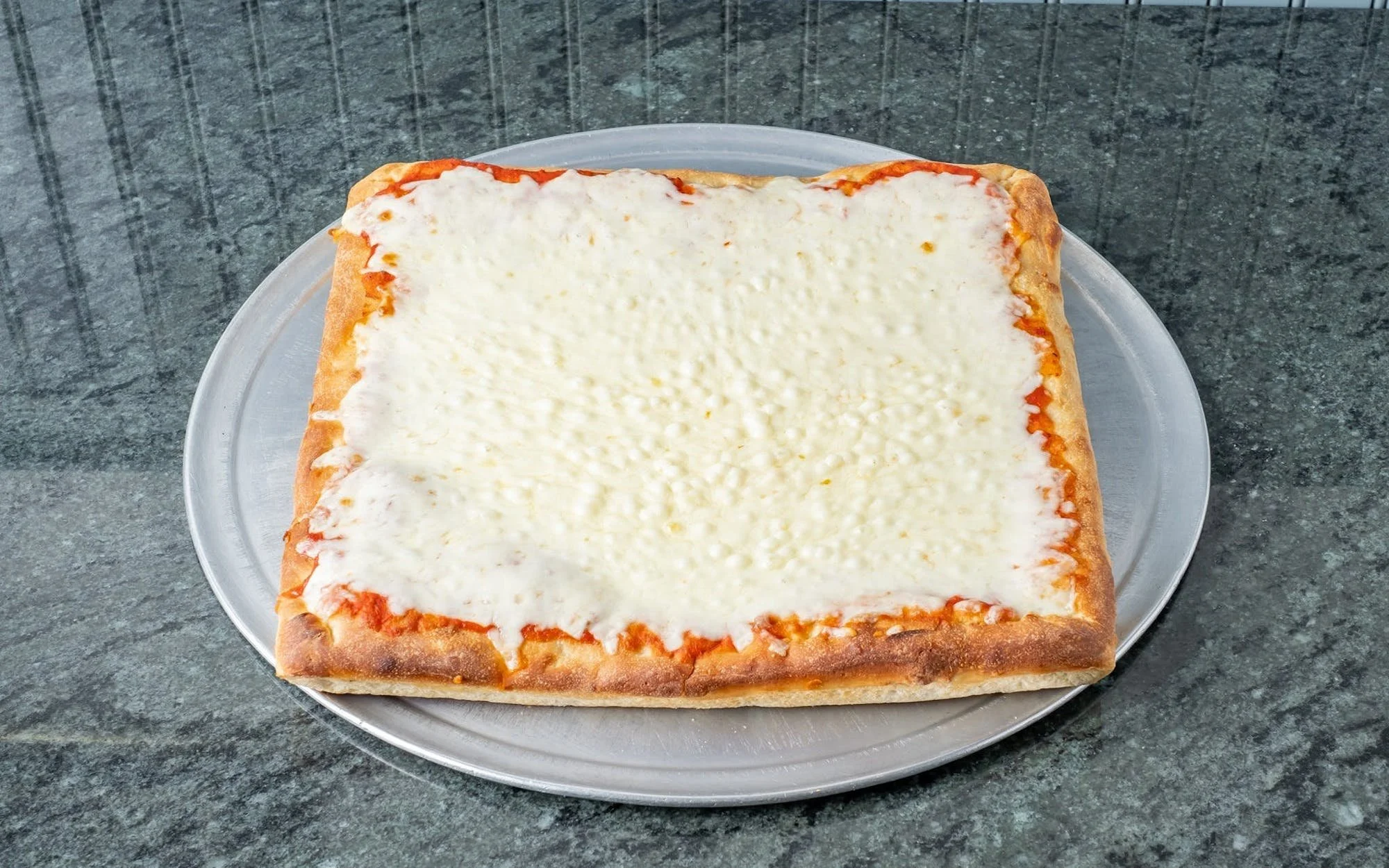Square slice of cheese pizza on a clear glass plate on a dark gray countertop.