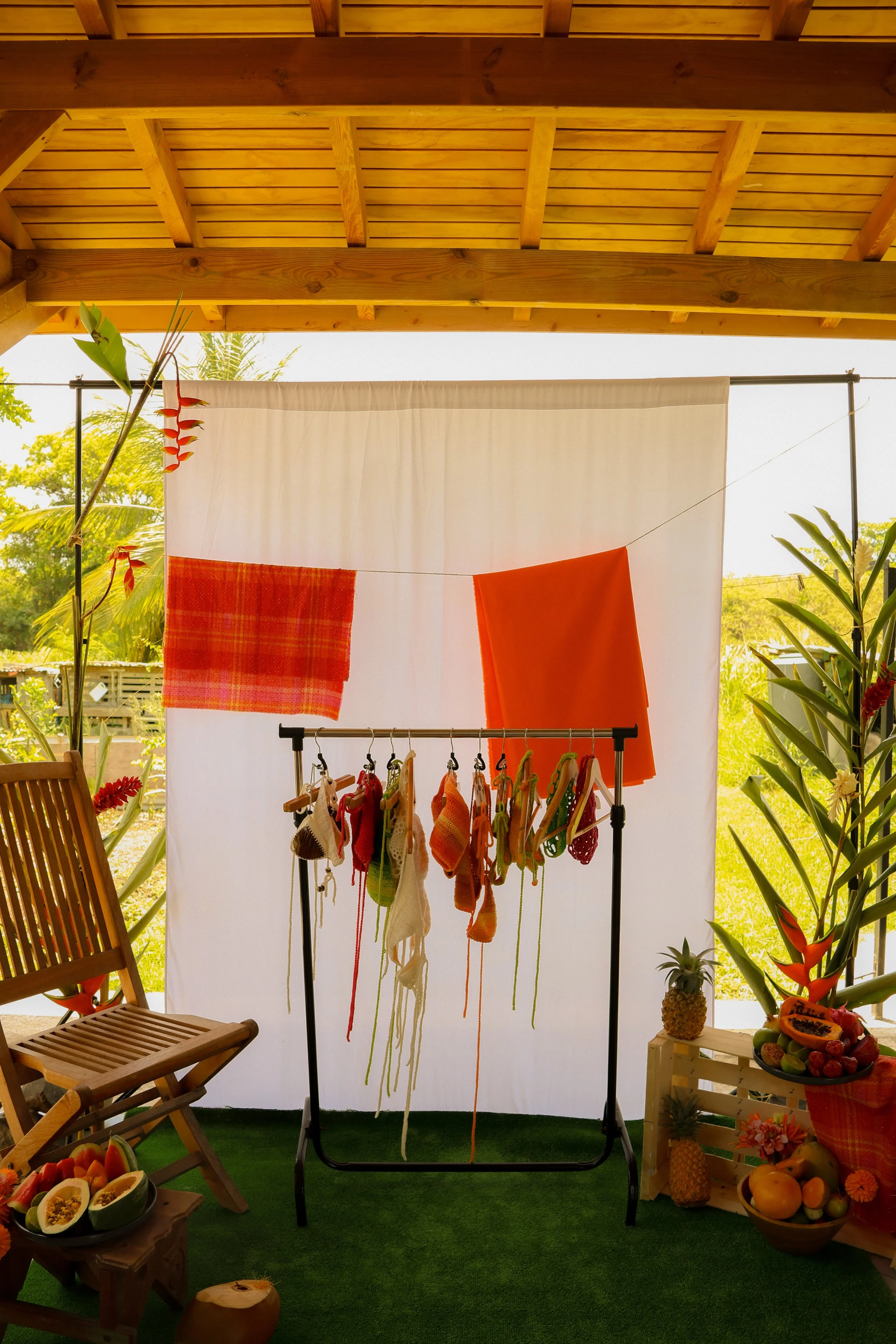 Balcon avec une tenture blanche derrière, vêtements tricotés suspendus, fruits frais, et décorations tropicales