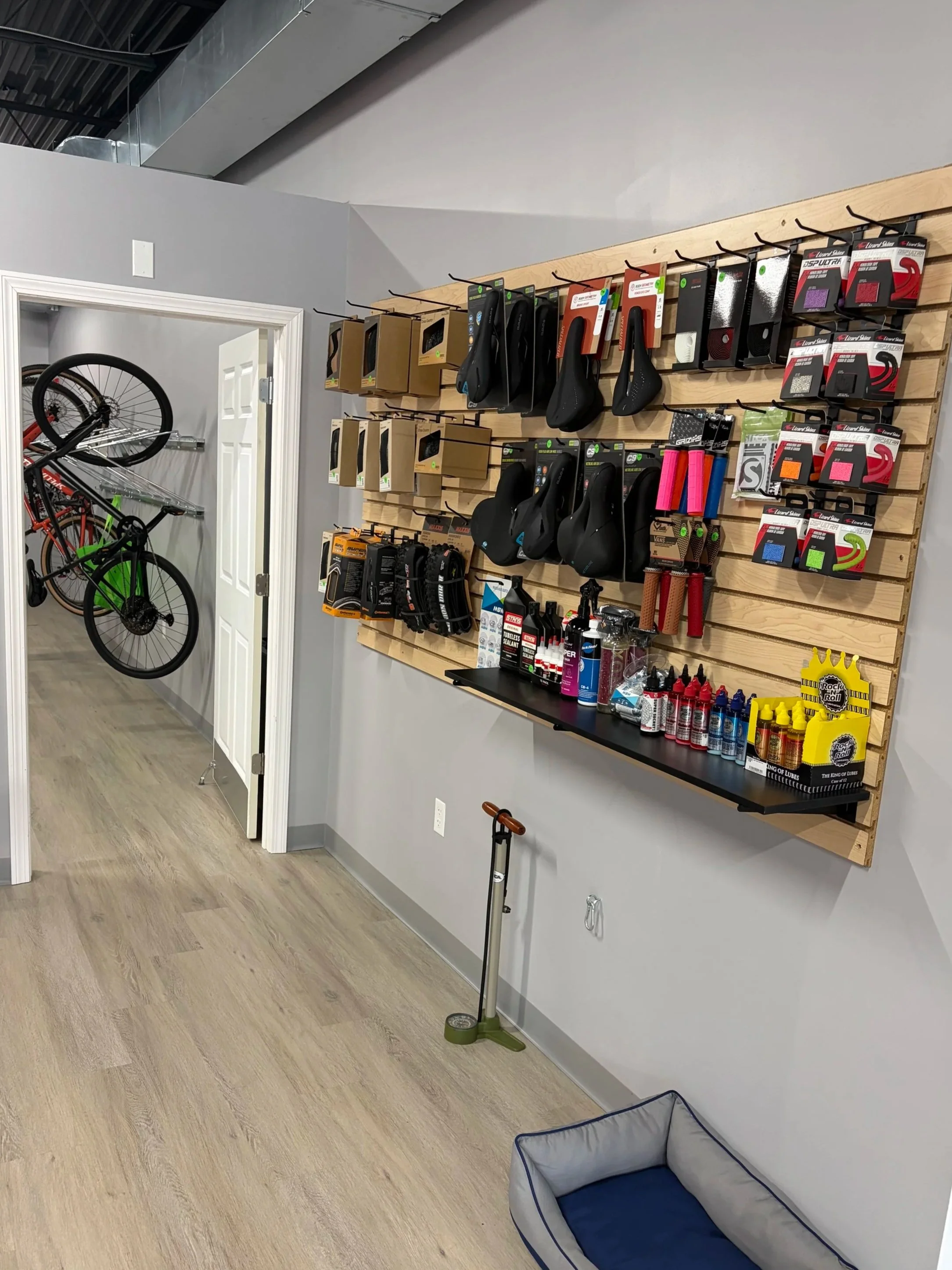 Bike shop interior displaying bicycle saddles, handlebars, grips, tires, tools, lubricants, and accessories on a wooden wall, with bicycles visible through the doorway.