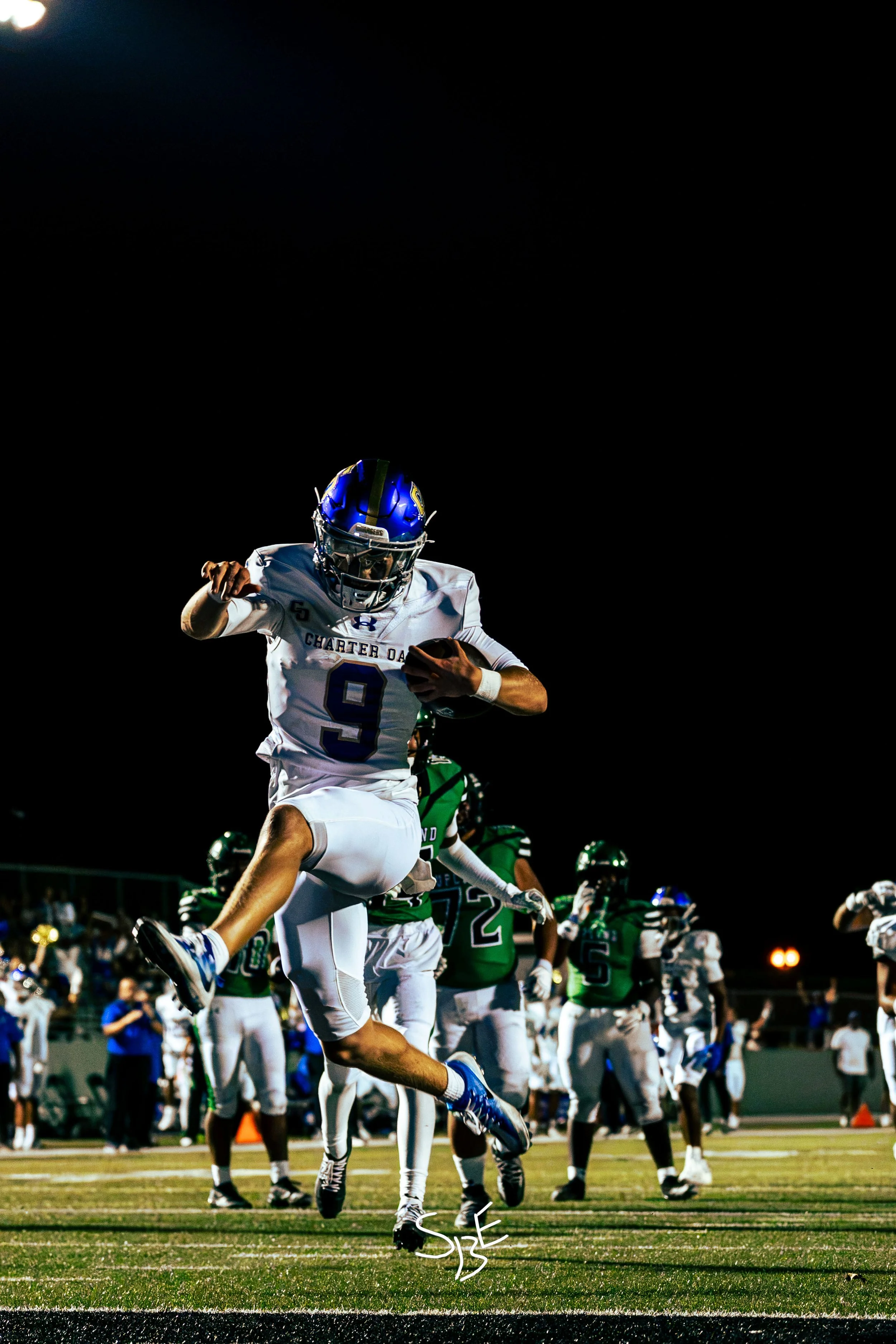 A football player wearing a white jersey and blue helmet runs with the football during a game at night. Other players and spectators are visible in the background.