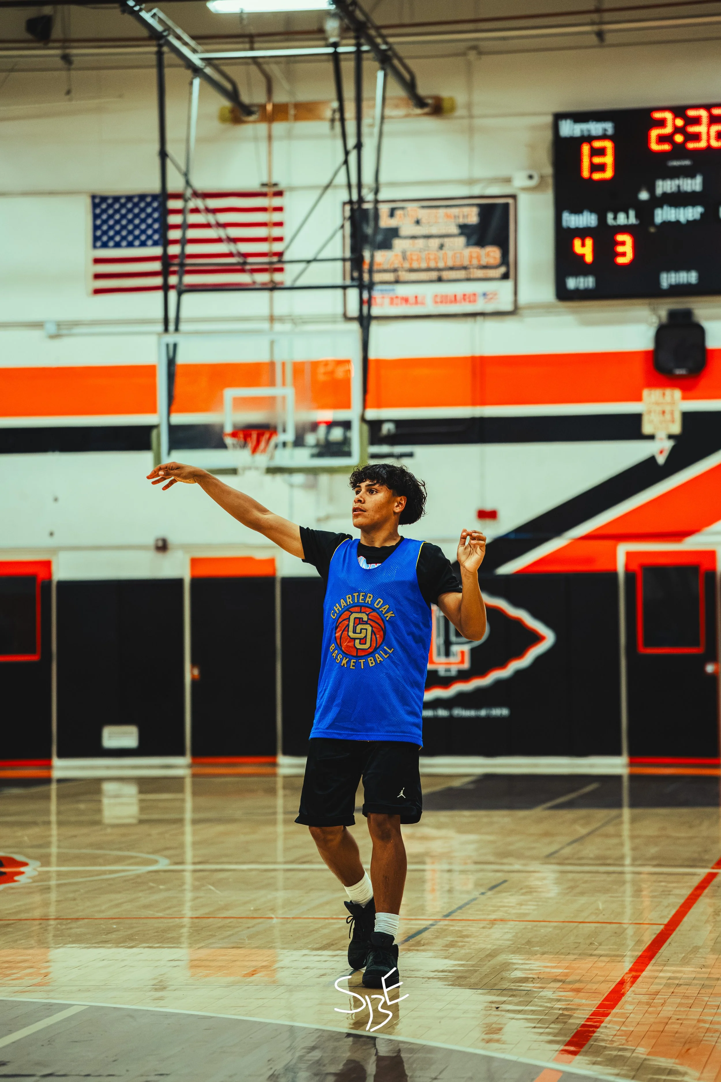 A young male basketball player practicing shooting in an indoor gymnasium, wearing a blue jersey with a logo, black shorts, and black sneakers, with an American flag hanging on the wall and a scoreboard in the background.