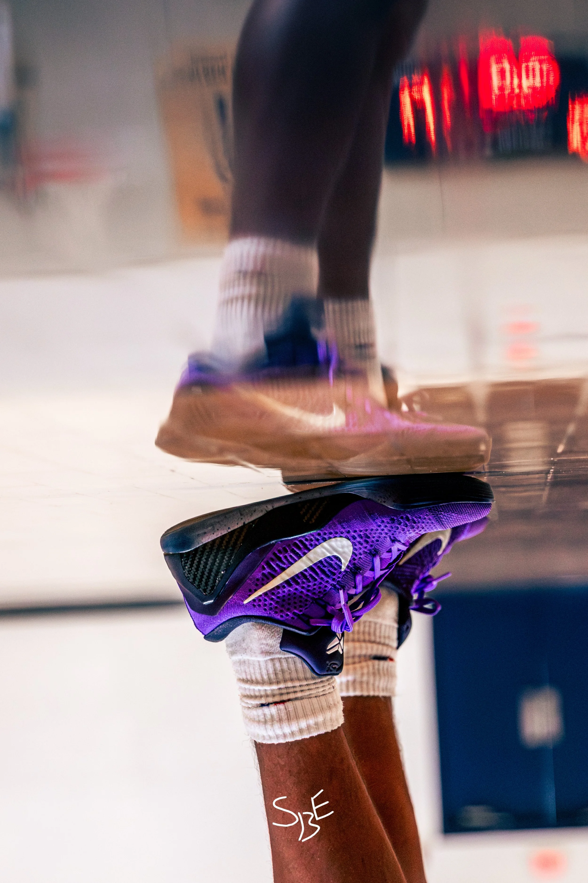 Close-up of a person wearing purple Nike running shoes, striped socks, beige shorts, and a knee brace, standing on a reflective gym floor.