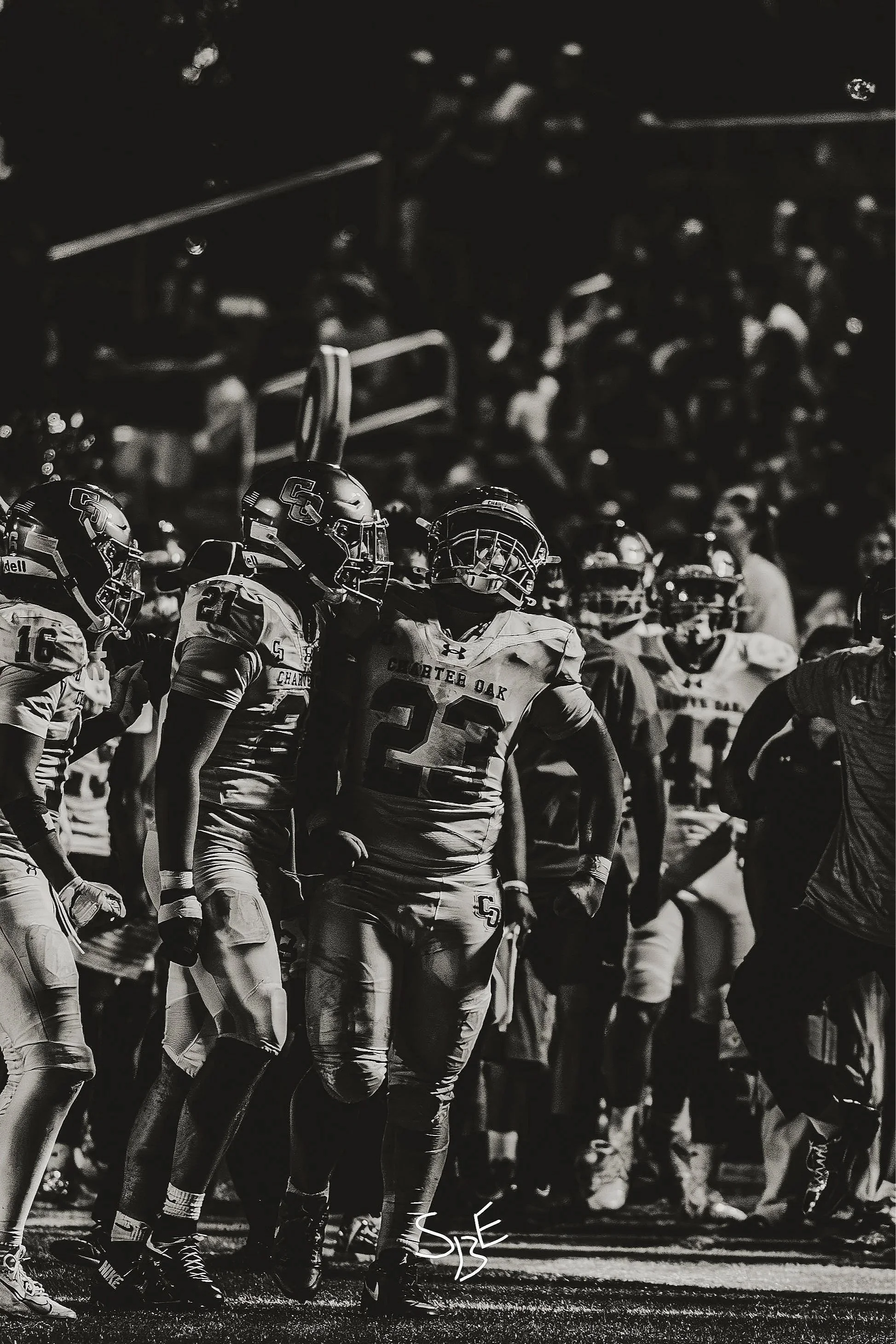 Black and white photo of a group of football players standing on the field during a game, wearing uniforms with helmets and pads, with some players in motion and others standing still.