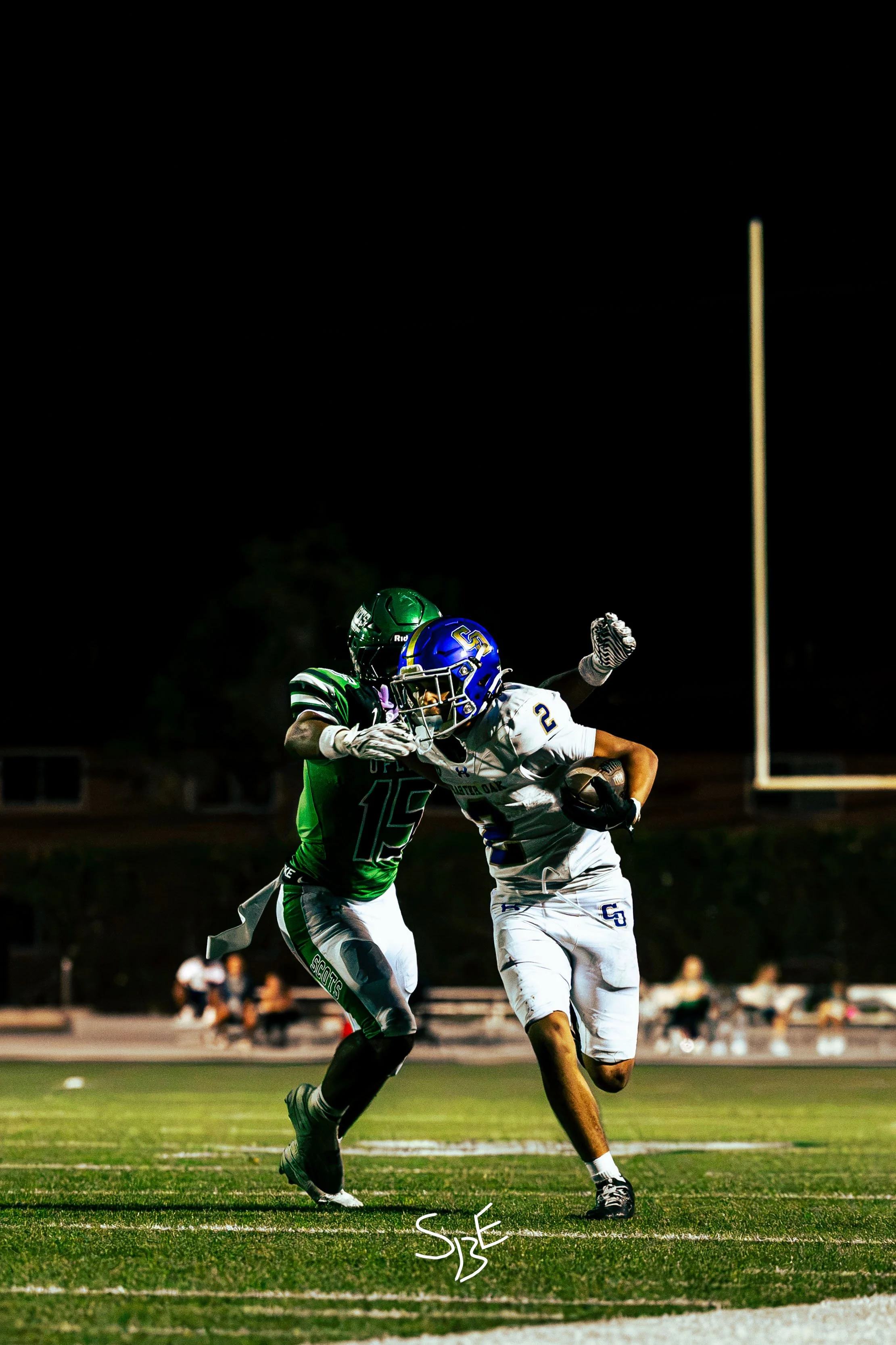 Two football players in action during a game at night, with one player from the team in white and the other from the team in green. The player in white is carrying the ball and trying to evade the defender in green. The scene is illuminated by stadiu