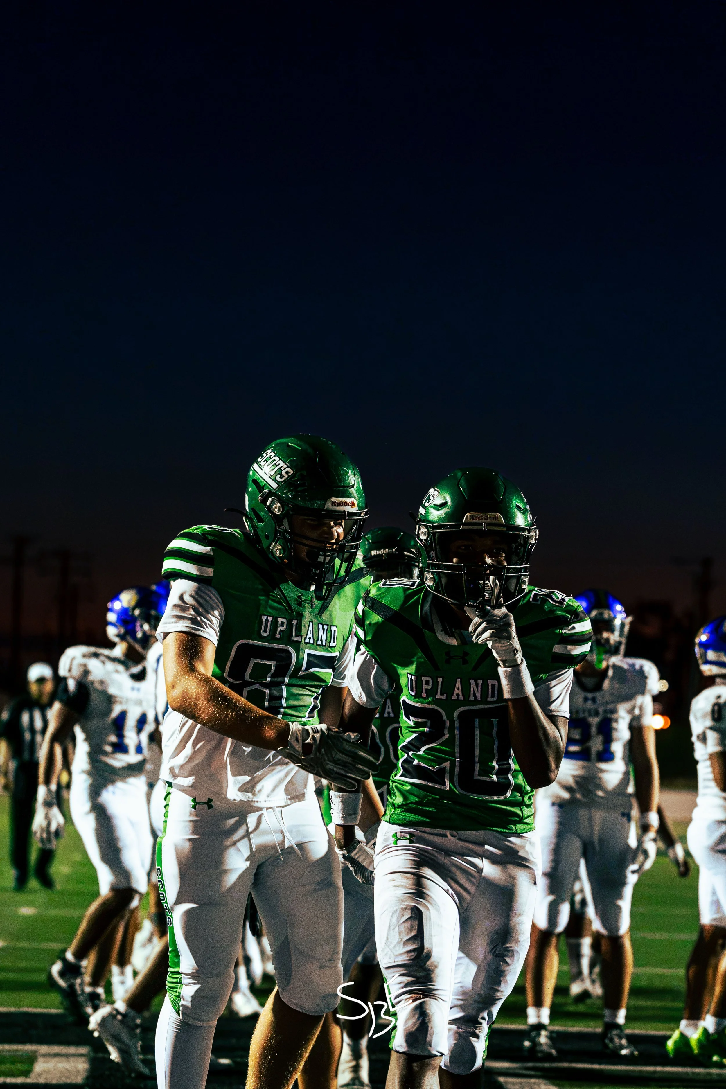 High school football players in green and white uniforms standing on the field at night, some with helmets on, during a game or practice.