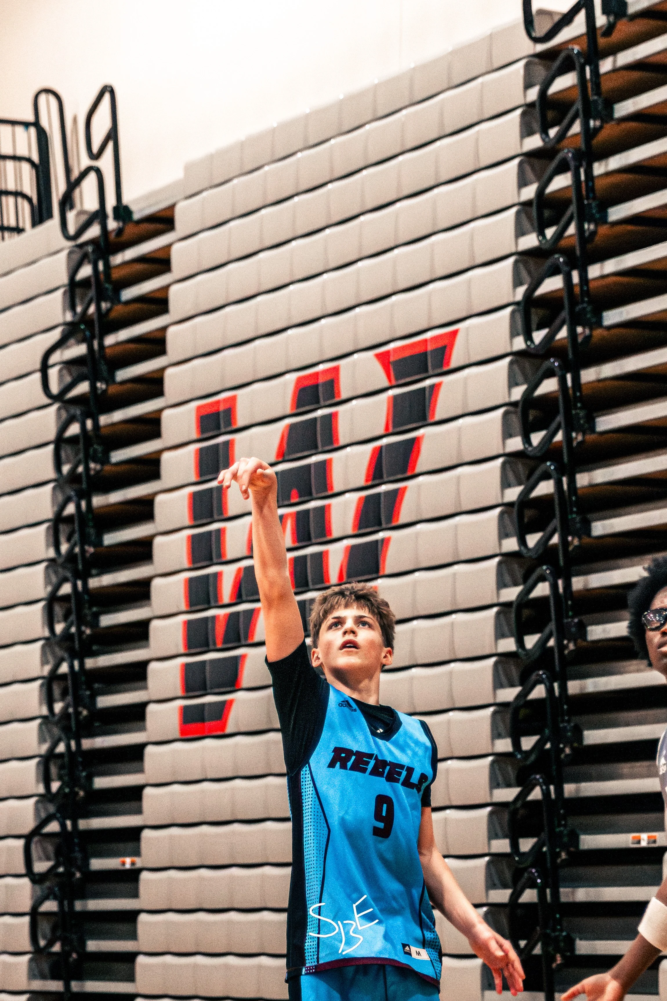 A young basketball player in a blue uniform with the number 9, raising his right arm during a game or practice in an indoor gym with a basketball court.