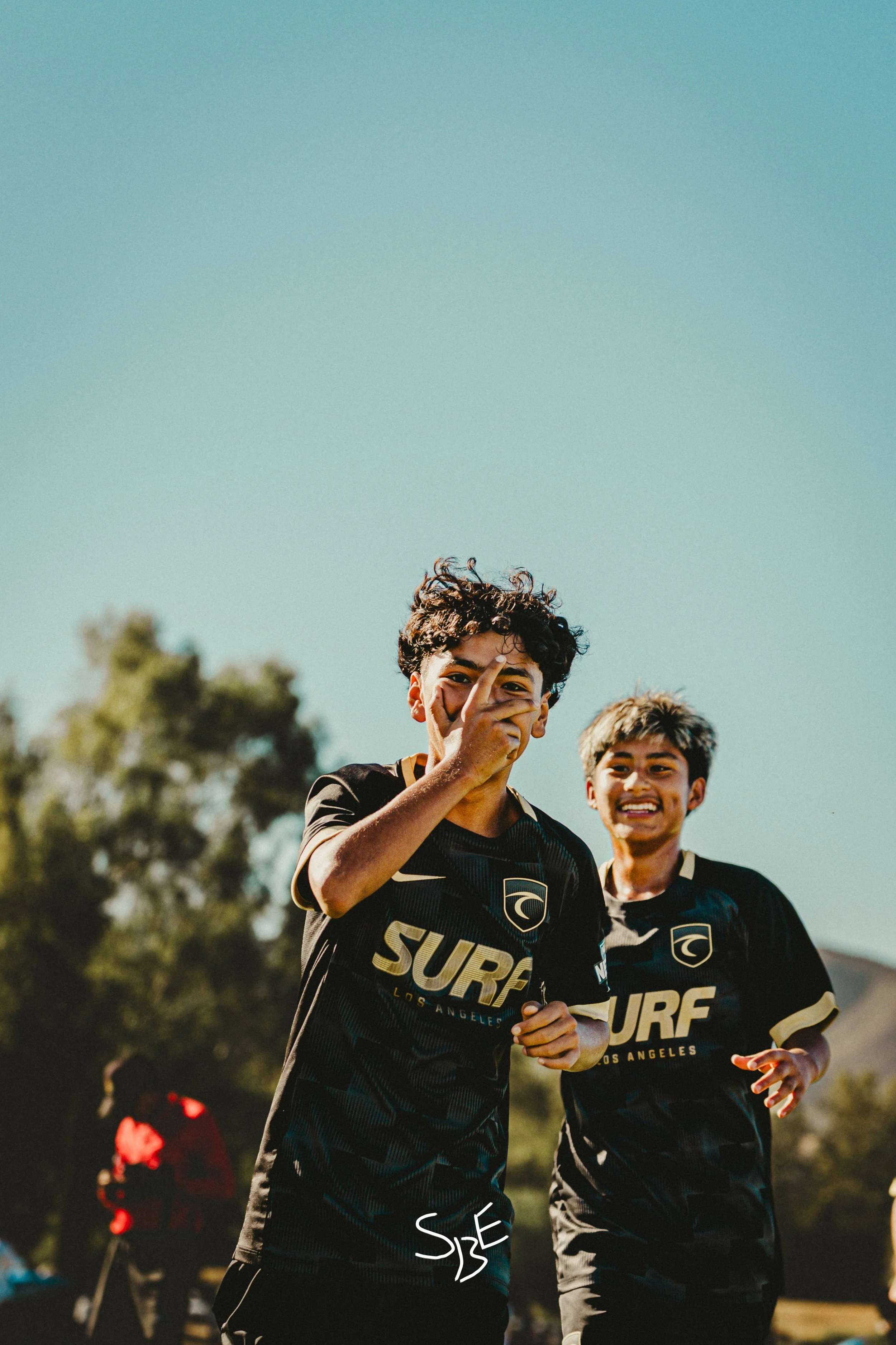 Two young male soccer players with athletic builds wearing black jerseys with gold accents and the word 'SURF' on the front, smiling and celebrating on a soccer field under a clear blue sky.