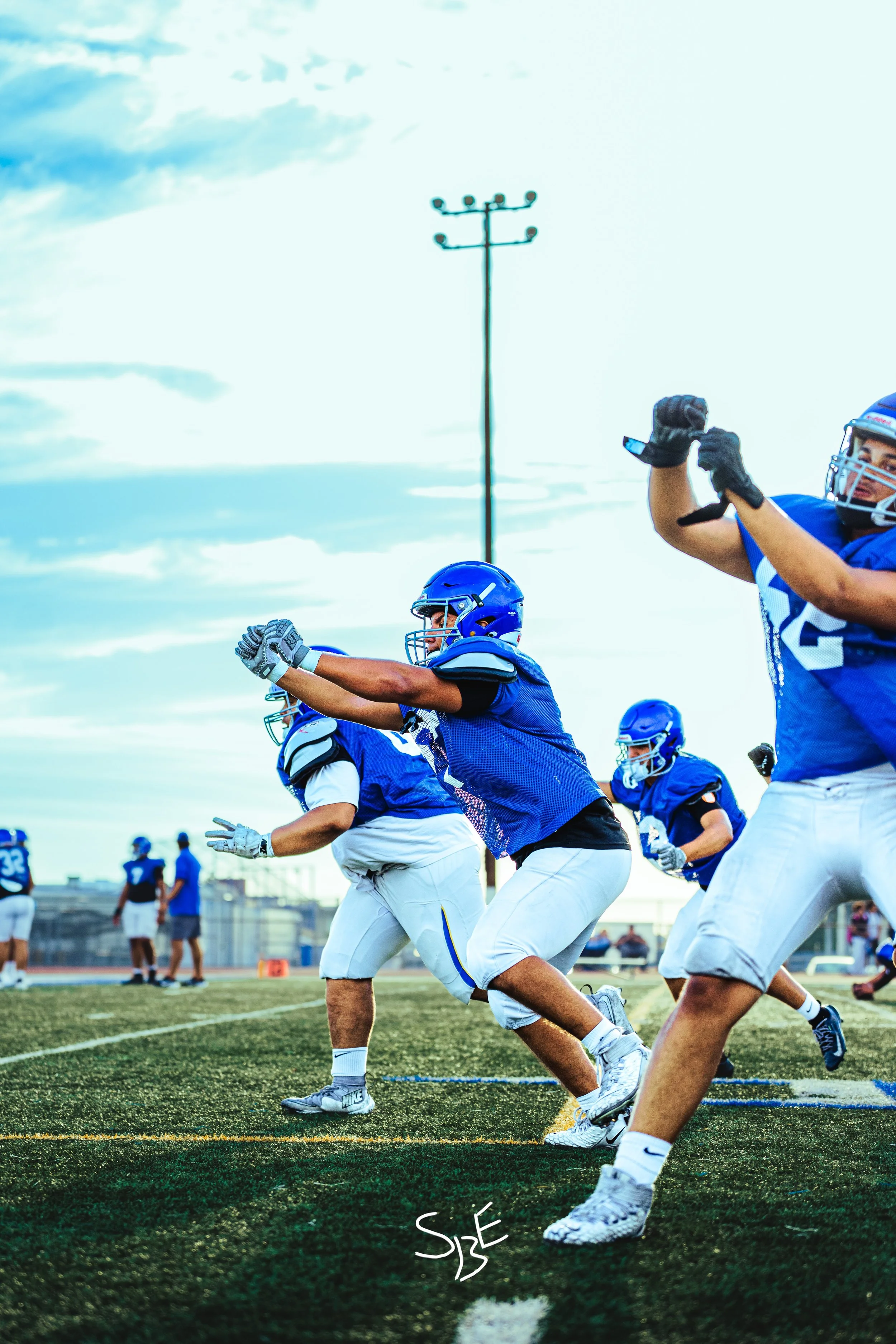Football players in blue and white uniforms practice running drills on a football field at sunset during a game or practice session.