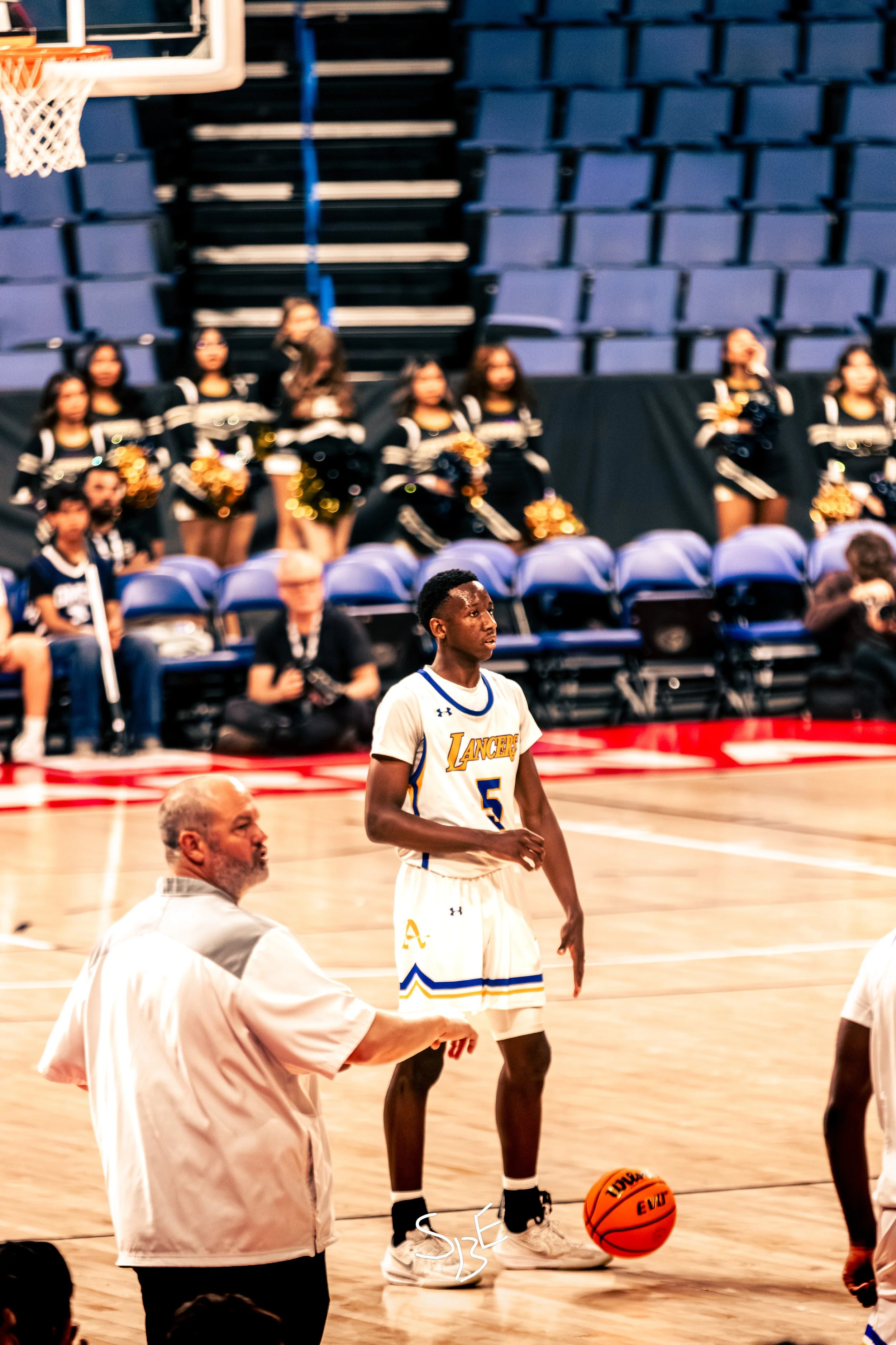 A basketball player in a white and blue uniform, with the number 5, stands on the court holding a basketball. An older man in a white shirt gestures towards the player. In the background, cheerleaders with pom-poms and a basketball hoop are visible, 