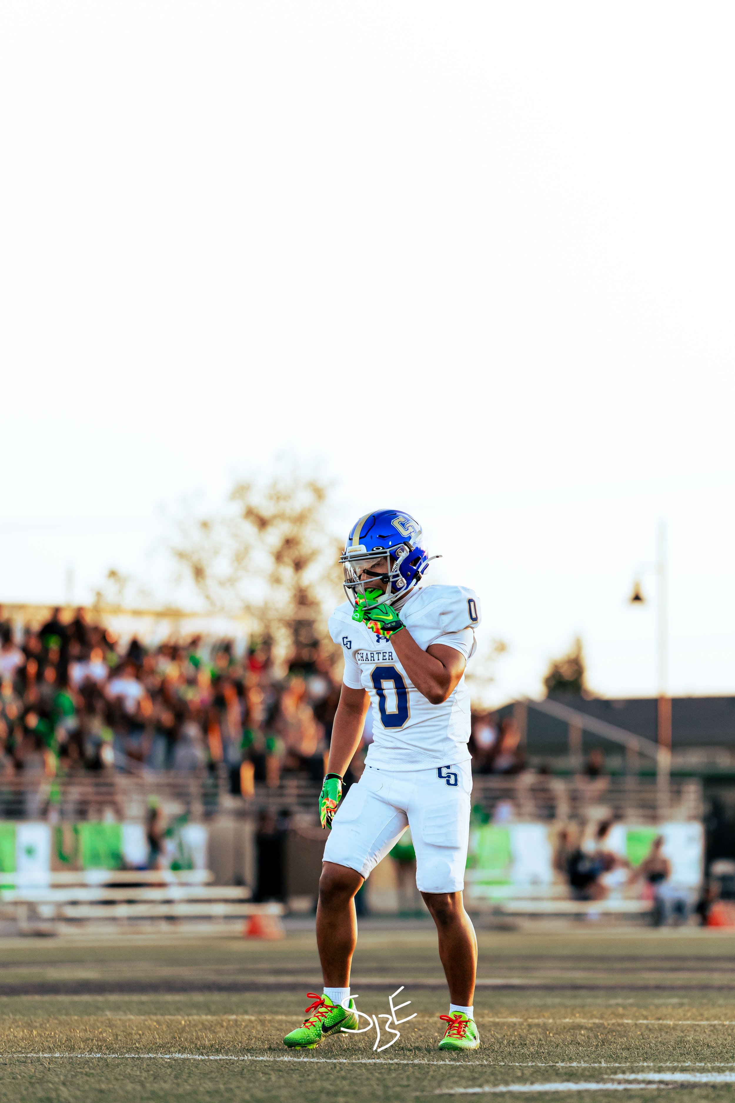 A football player wearing a white uniform with the name 'Charter' and the number 0, standing on a field during a game with spectators in the background. The player is adjusting a bright green helmet with red laces.