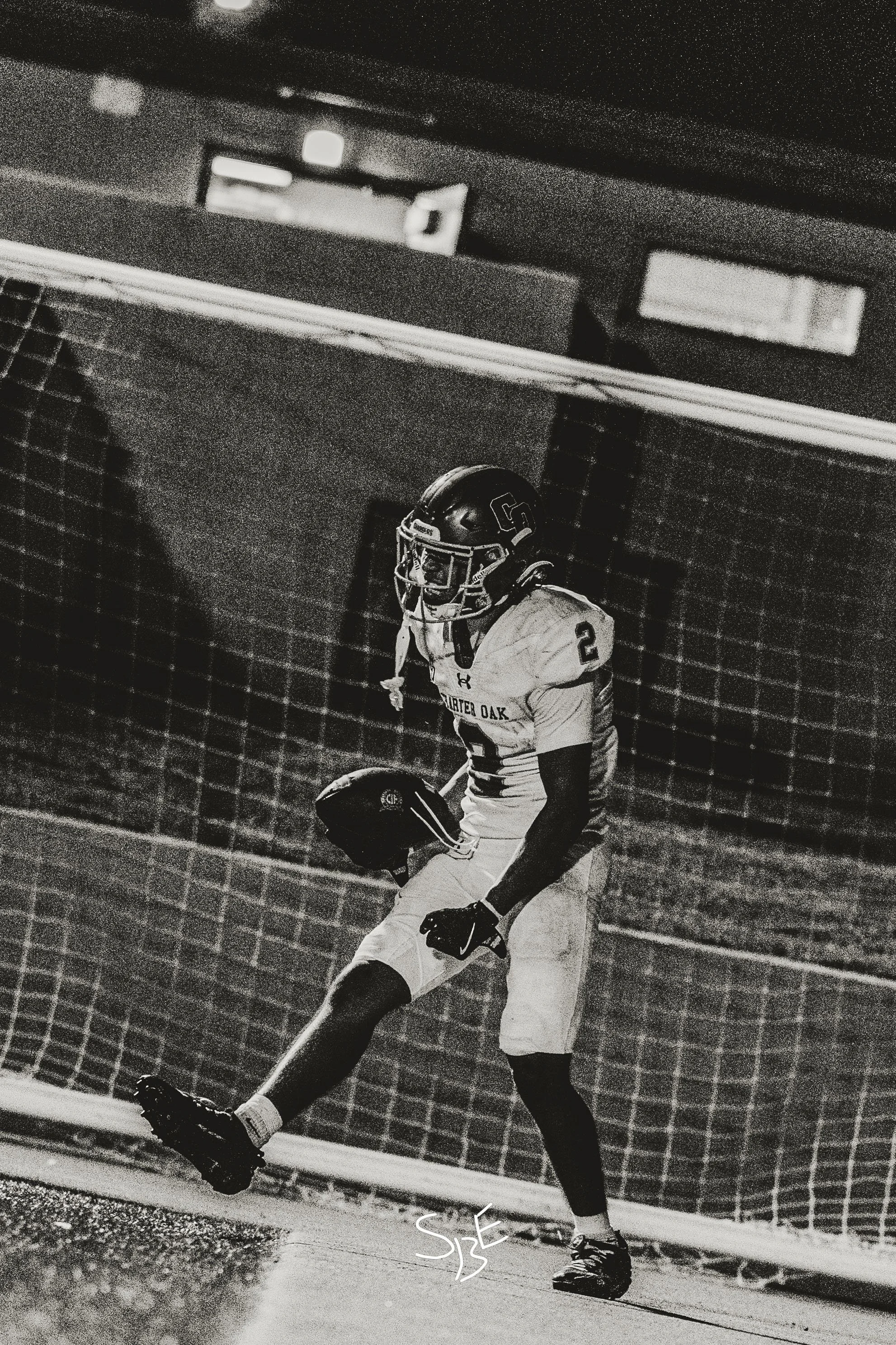 Black and white photograph of an American football player walking along the sideline at the stadium, holding a helmet in one hand, wearing a uniform with the number 2, and a helmet with a faceguard. The background shows a chain-link fence and some si