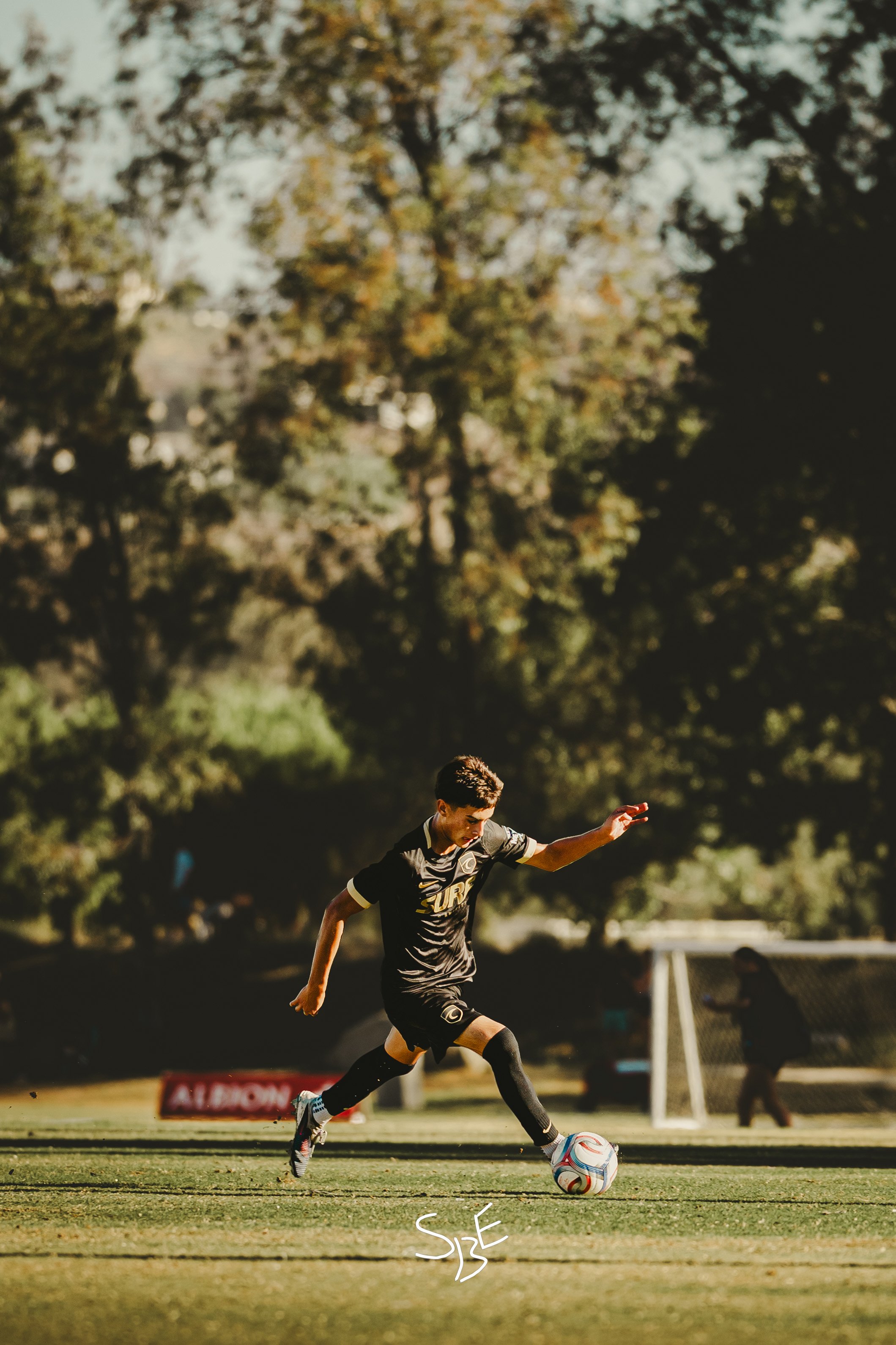 A young male soccer player in black uniform kicking a soccer ball on an outdoor field with trees in the background during daytime.