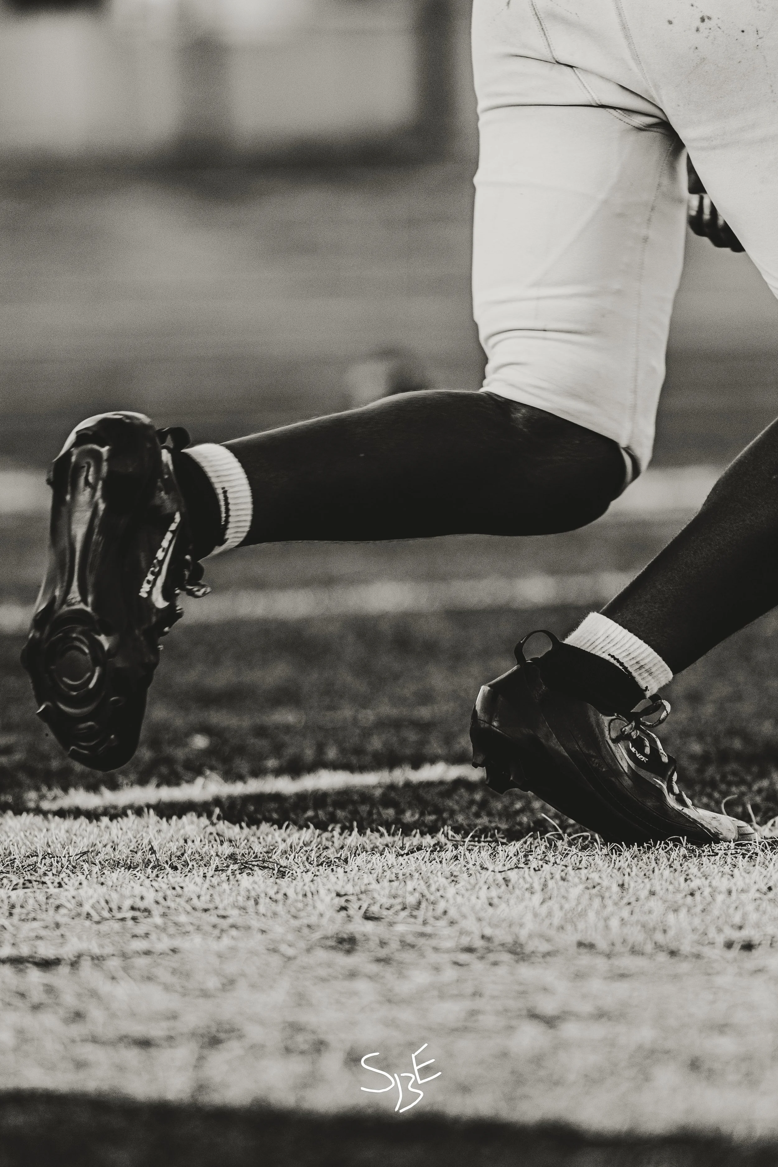 A person wearing athletic shoes and socks is in a crouched position on a grassy field, with their arm extended downward. The photo is in black and white and focuses on the lower body and shoes.