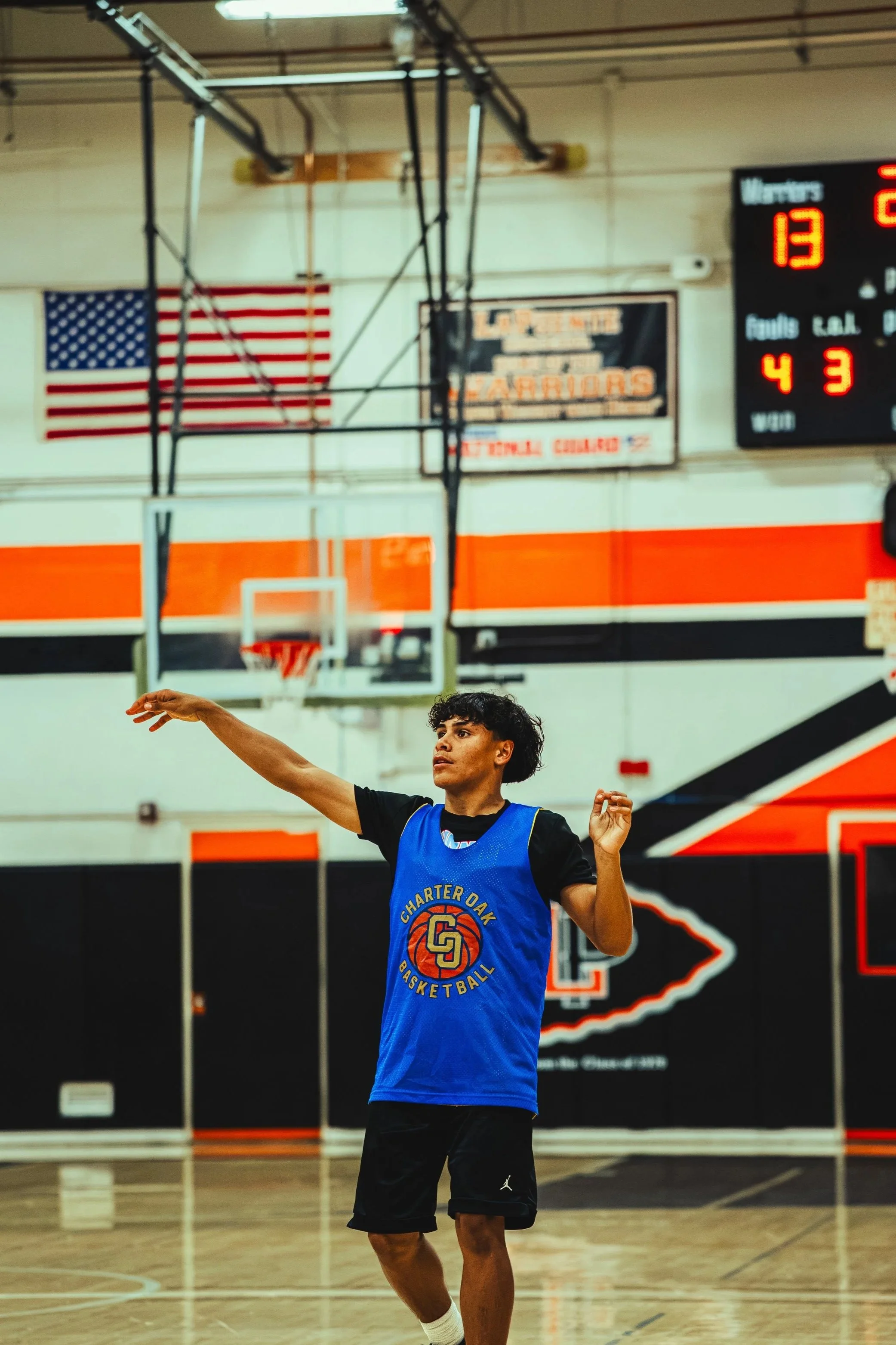 A young male basketball player with curly dark hair practicing free throws on an indoor basketball court. He is wearing a blue jersey with sports logos and black shorts, with a focused expression and arms extended for the shot.