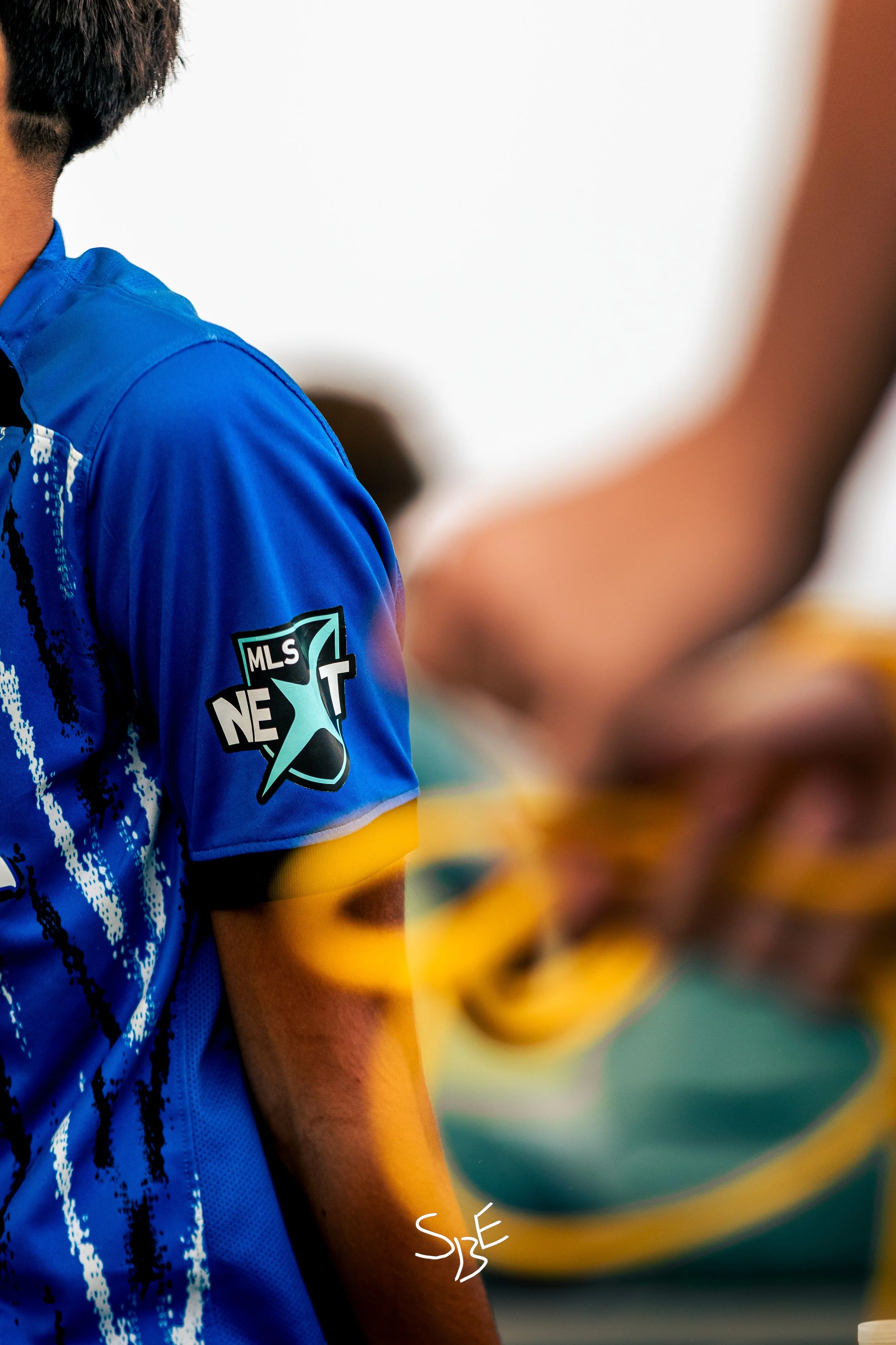 Close-up of a person wearing a blue sports jersey with a MLS Next badge, standing in a sports setting with blurred people in the background.