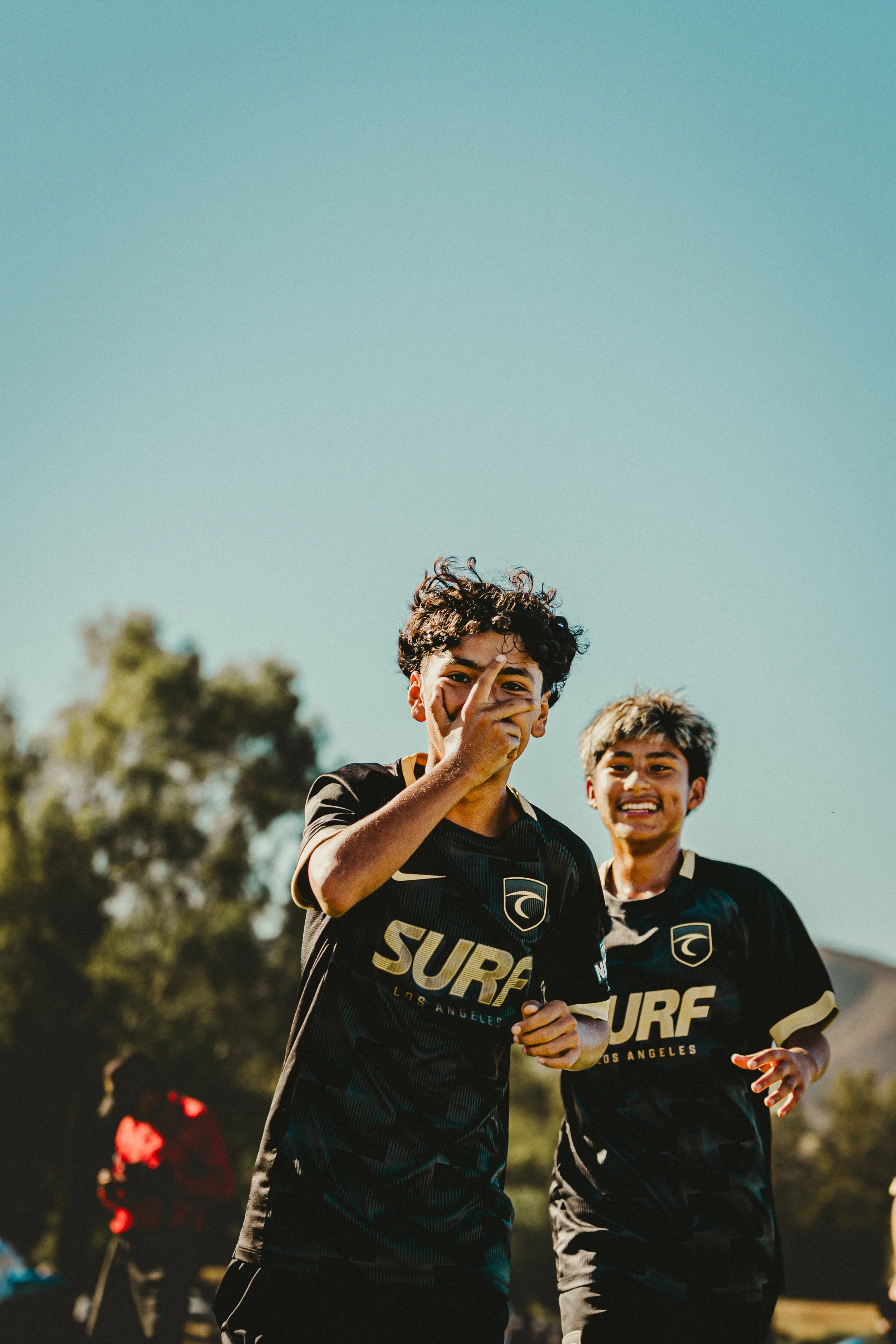 Two young male soccer players in black jerseys with gold and white accents, celebrating on the field during a game, with trees and a clear blue sky in the background.