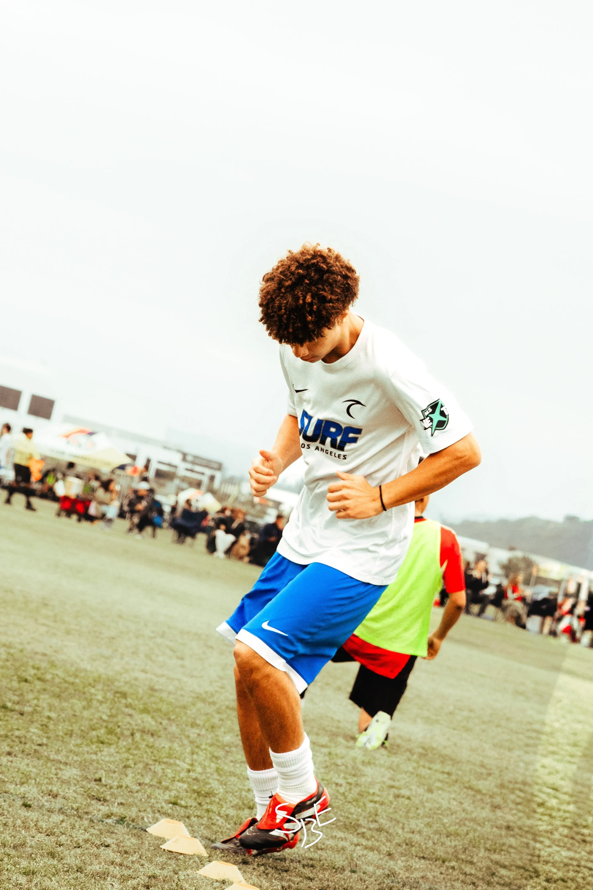 A young male soccer player in a white jersey and blue shorts practicing drills on a field. The background shows a crowd and tents, indicating an outdoor sporting event.