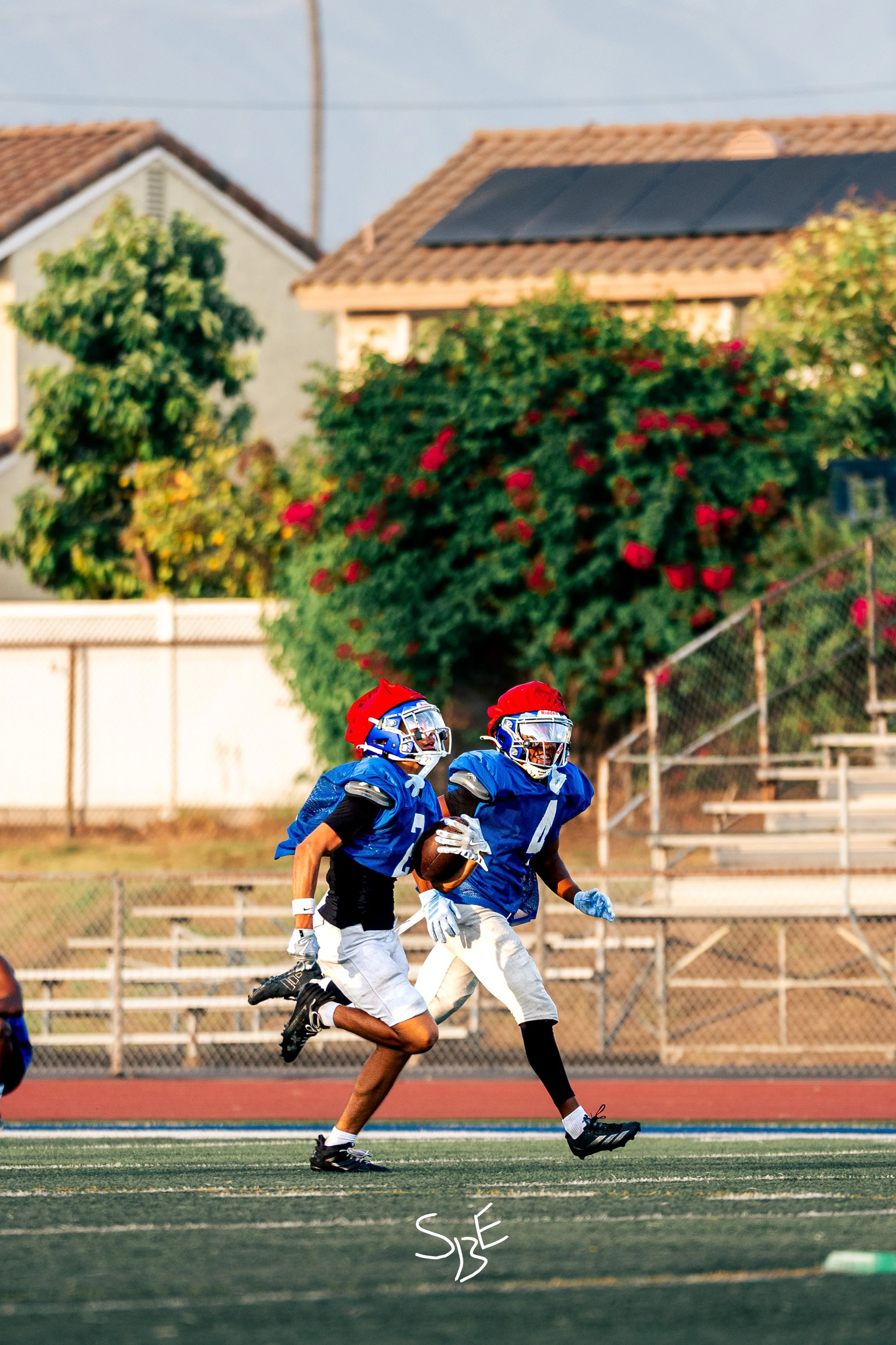 Two football players wearing blue jerseys and white pants running on an outdoor field during a game, with a chain-link fence, bleachers, trees, and houses in the background.