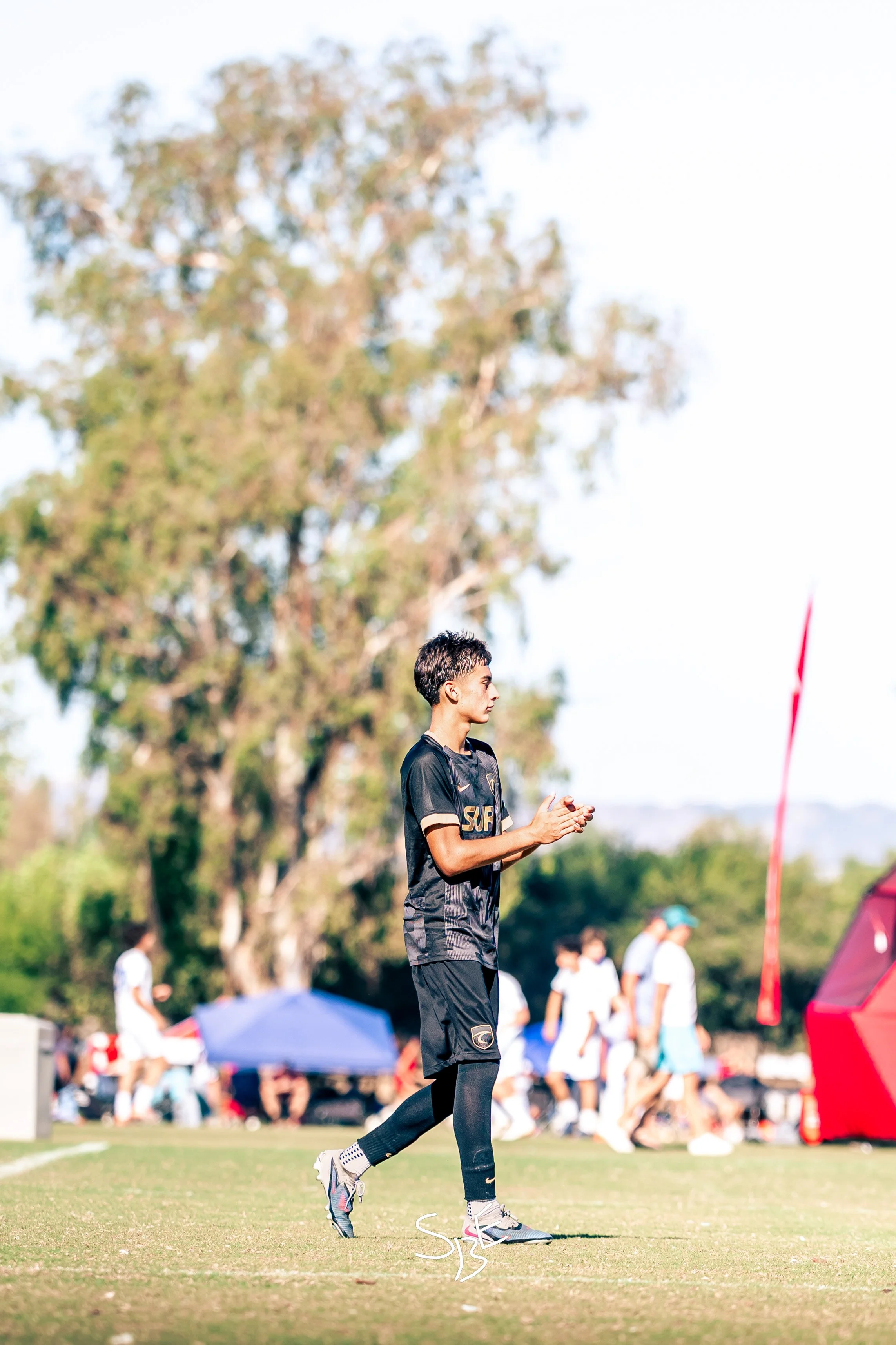 A young man, dressed in sportswear, running on a grassy field during a sporting event or practice. There are other people and tents in the background, with trees and a partly cloudy sky.
