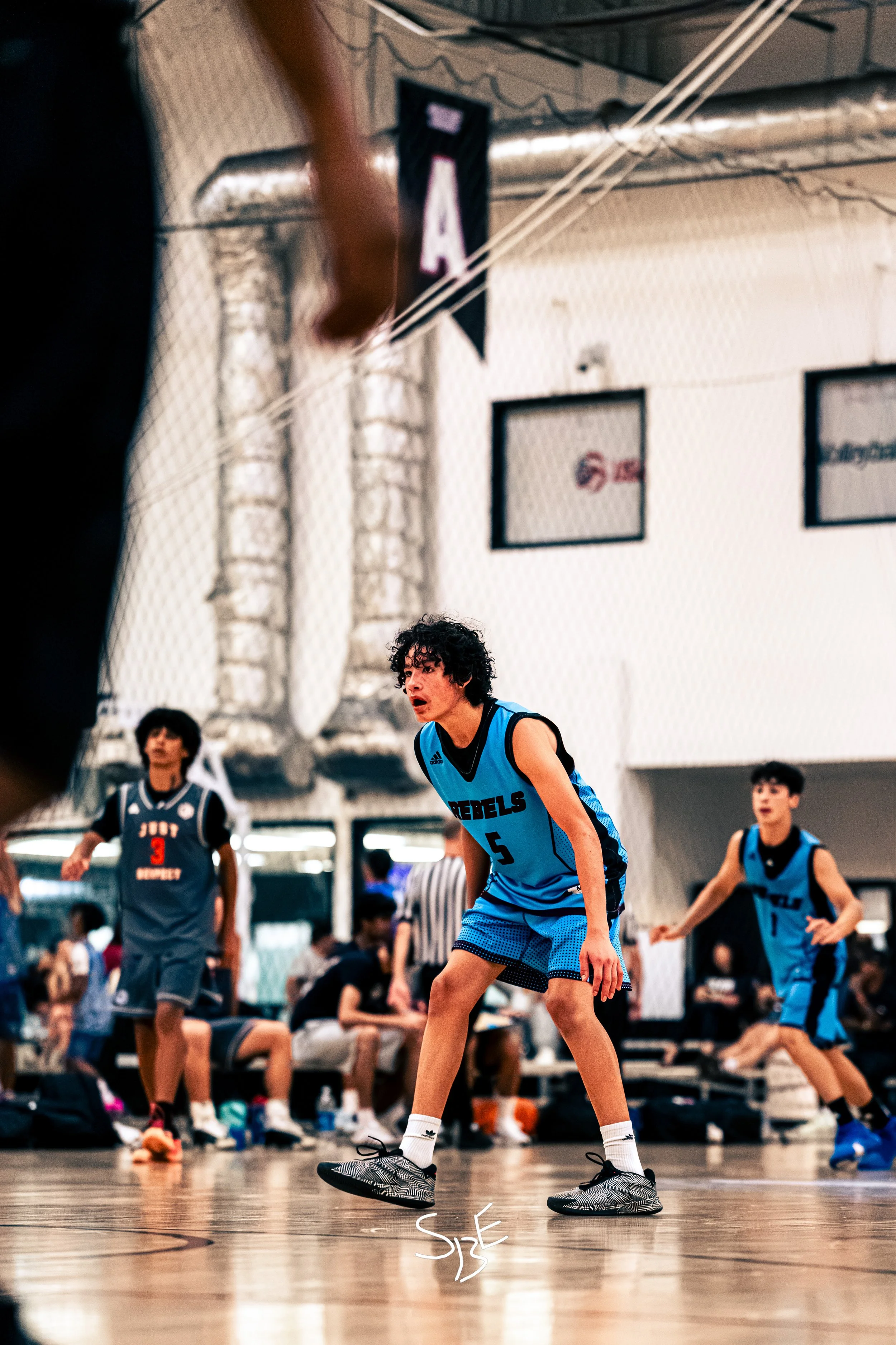Youth basketball game in progress with players wearing blue and black jerseys on an indoor court, a player in the foreground bent low, postured for defense, with other players in the background, some sitting and some standing.