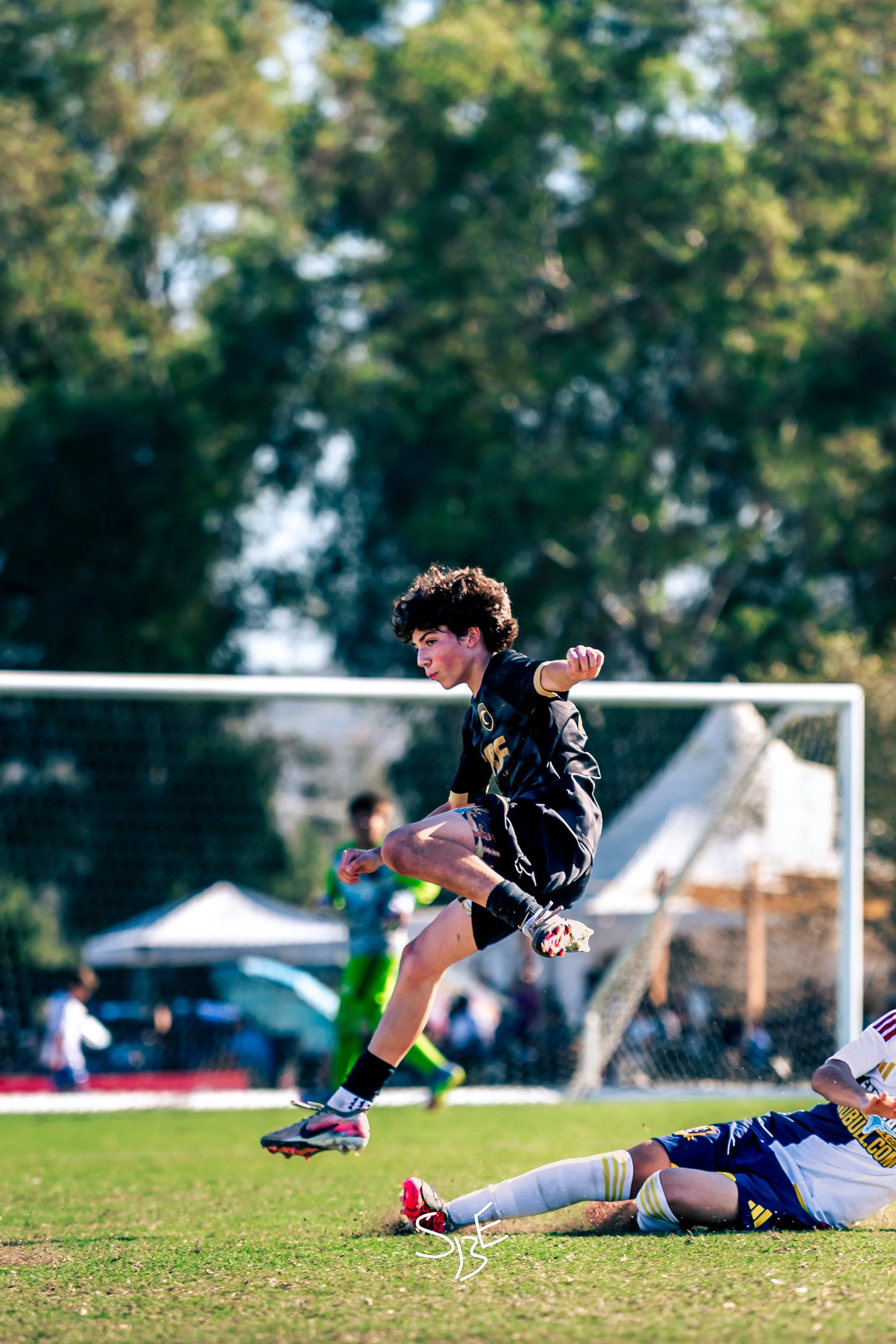 A soccer game where a player is mid-air, jumping over a fallen opponent on the field, with green trees and tents in the background.