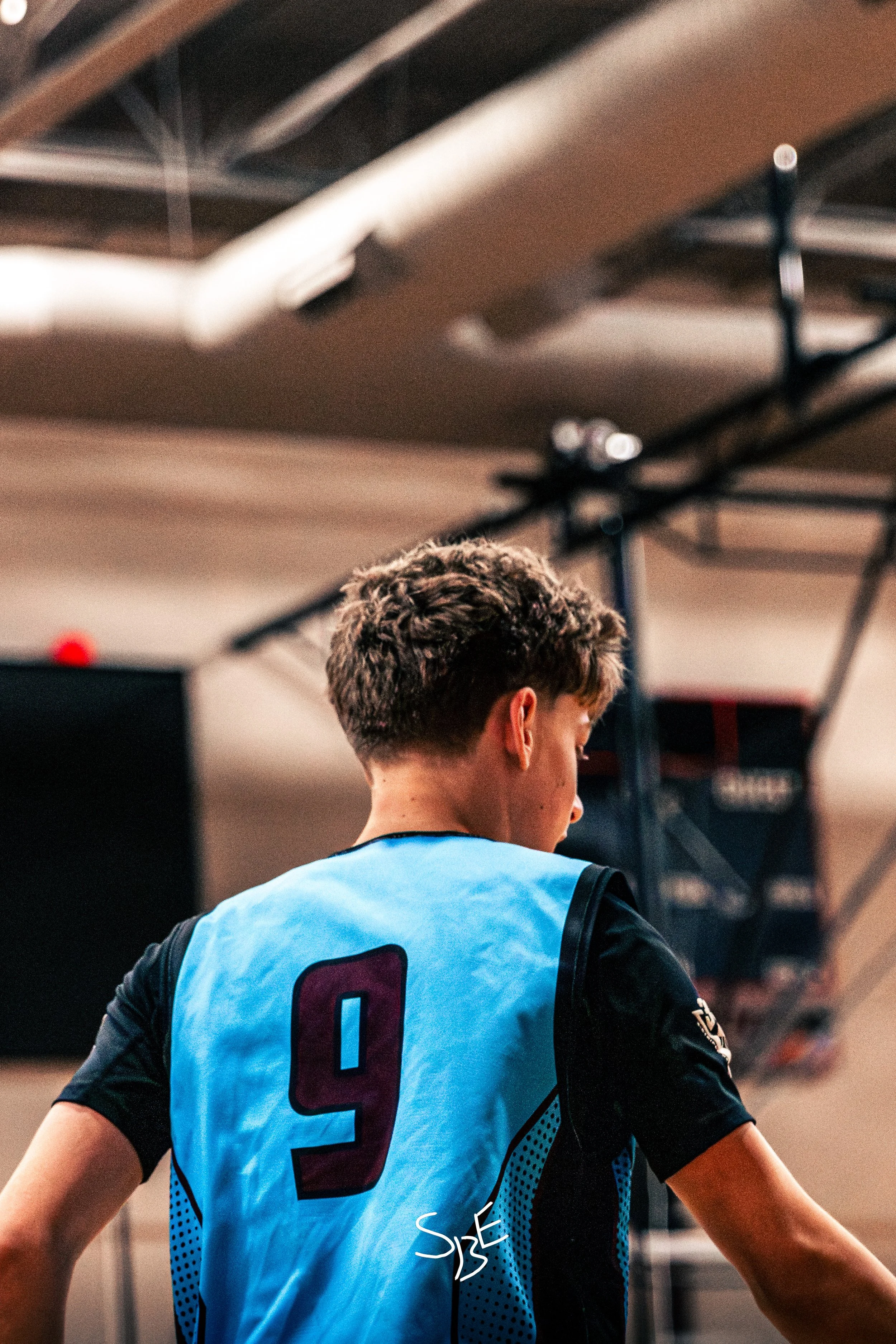 A person with short brown hair wearing a blue sports jersey with black sleeves and red number nine on the back, standing indoors.