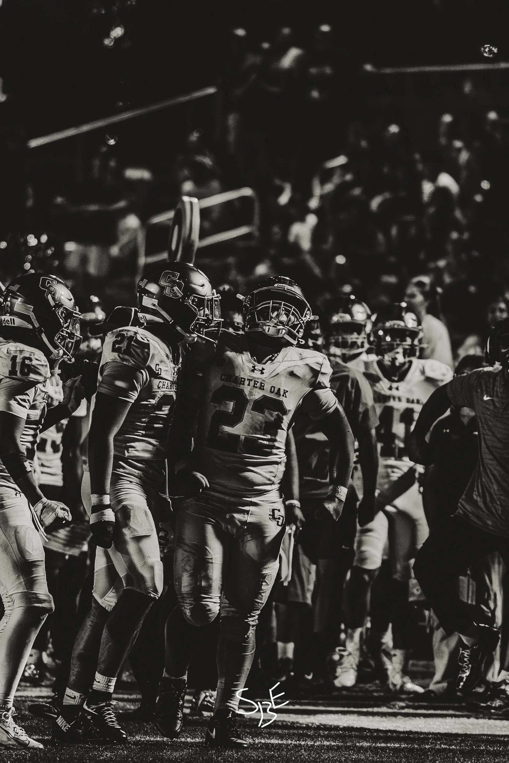 Black and white sports photograph of football players in helmets and jerseys during a game on the field, some players are standing and others are in motion.